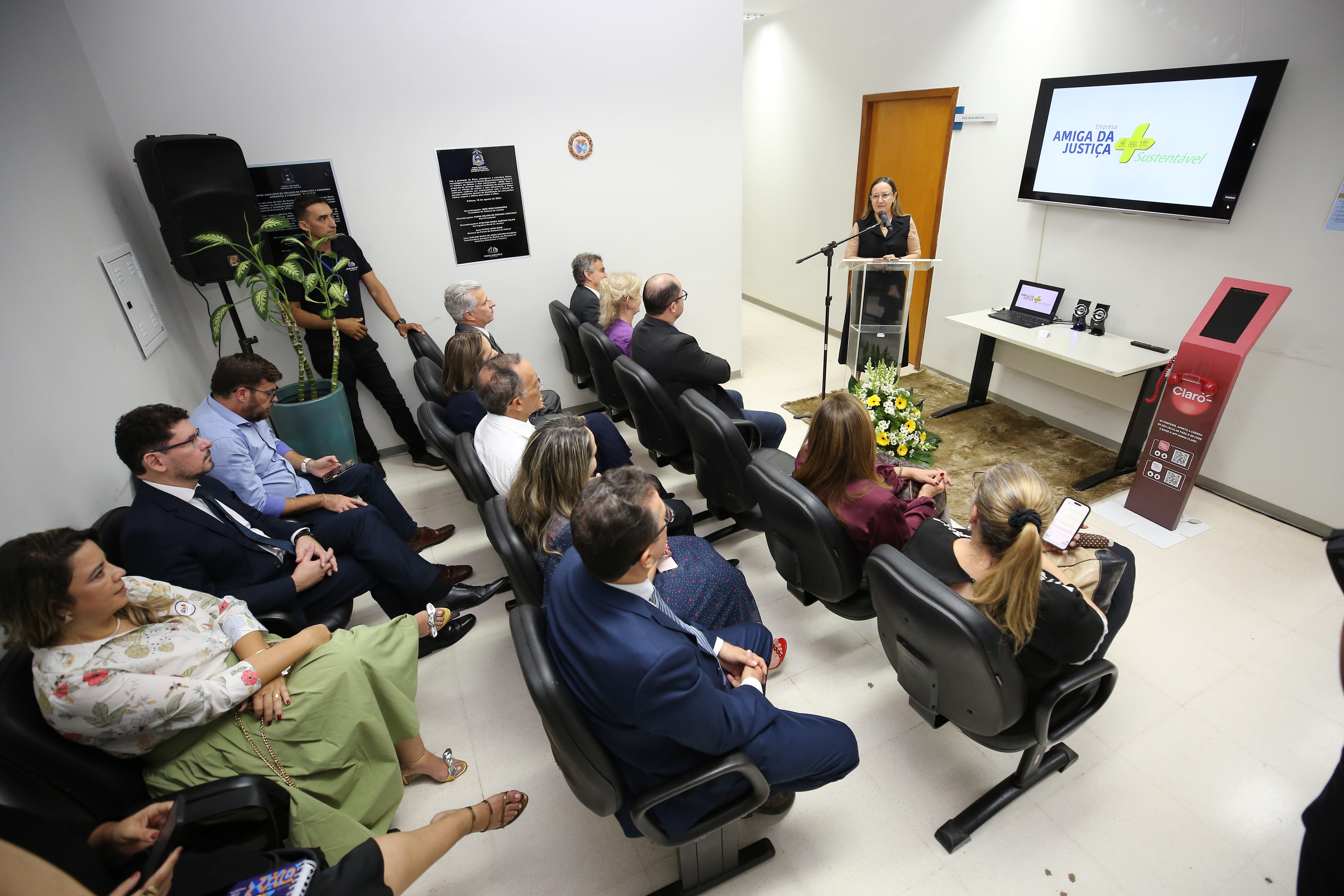 Solenidade com pessoas de costas sentadas numa pequena sala assistindo mulher falando ao púlpito. Atrás dela uma tela. 