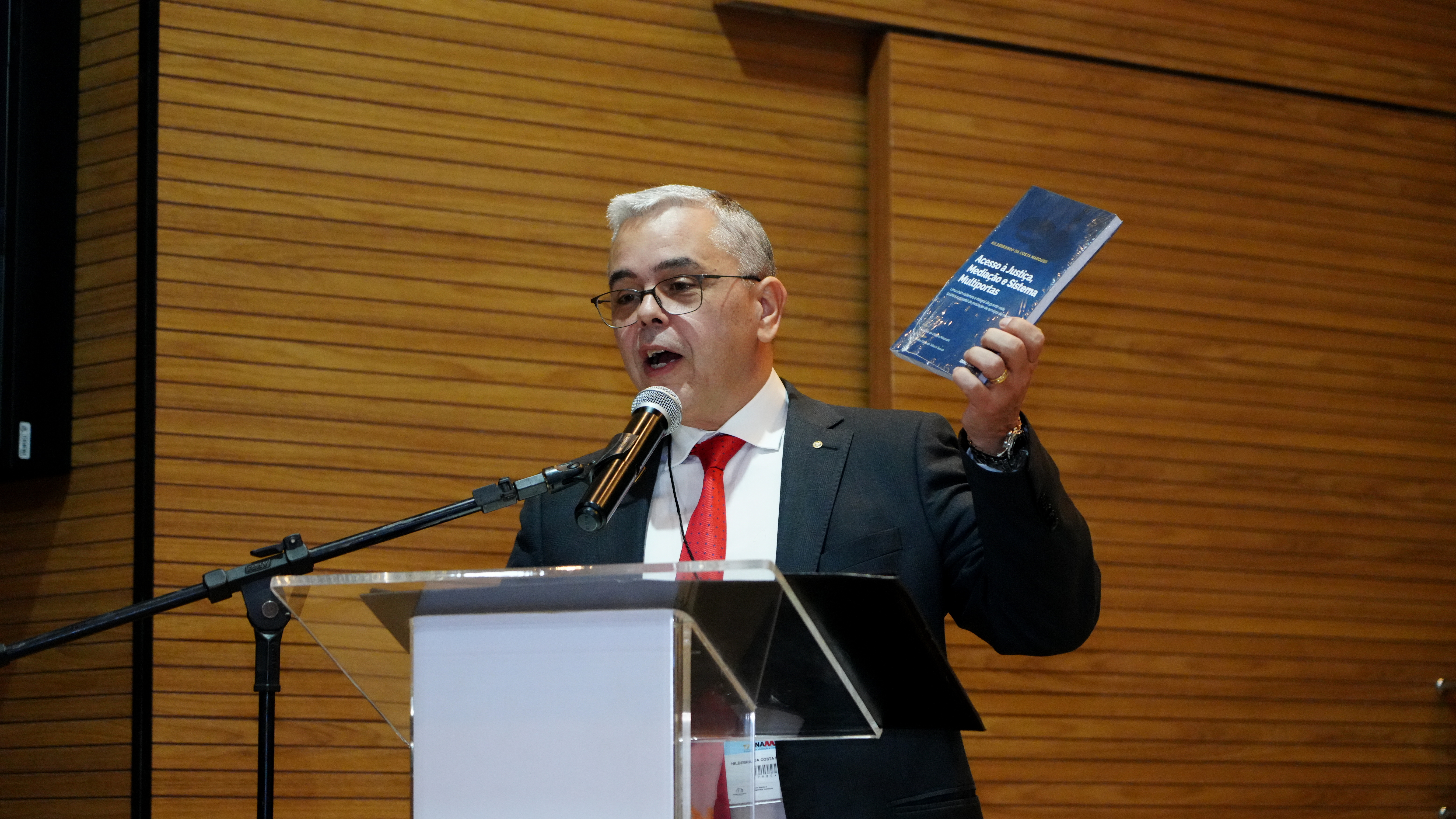 A imagem mostra um homem de meia-idade, vestido com terno e gravata, segurando um livro no palco do auditório do Tribunal de Justiça do Tocantins, durante o 17º Fórum Nacional de Mediação e Conciliação.