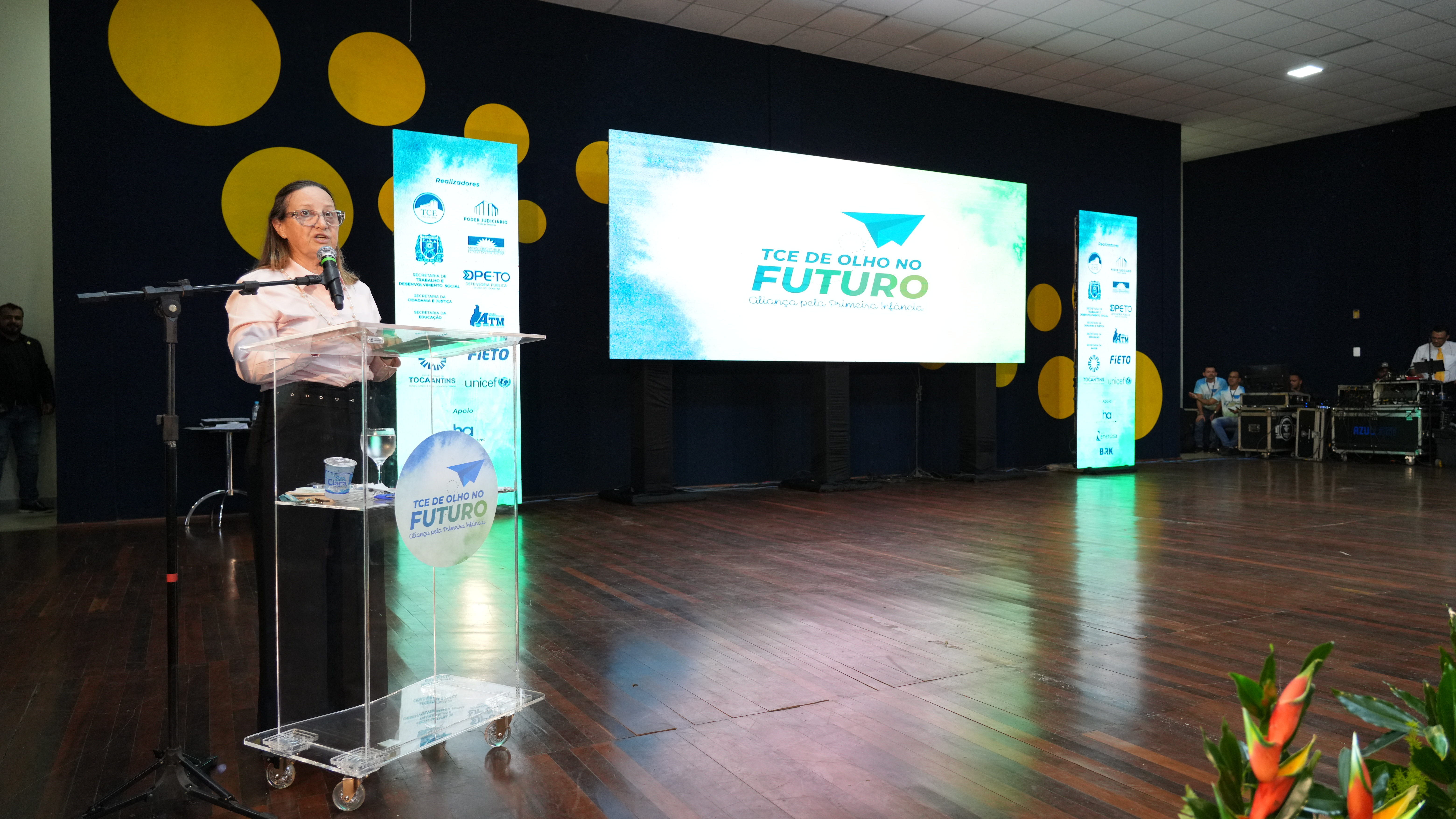 Mulher branca vestindo calça preta e camisa de manga comprida de cor rosa clara, fala ao púlpito em cima de um tablado. Ao fundo, uma tela projeta nome de projeto voltado à crianças