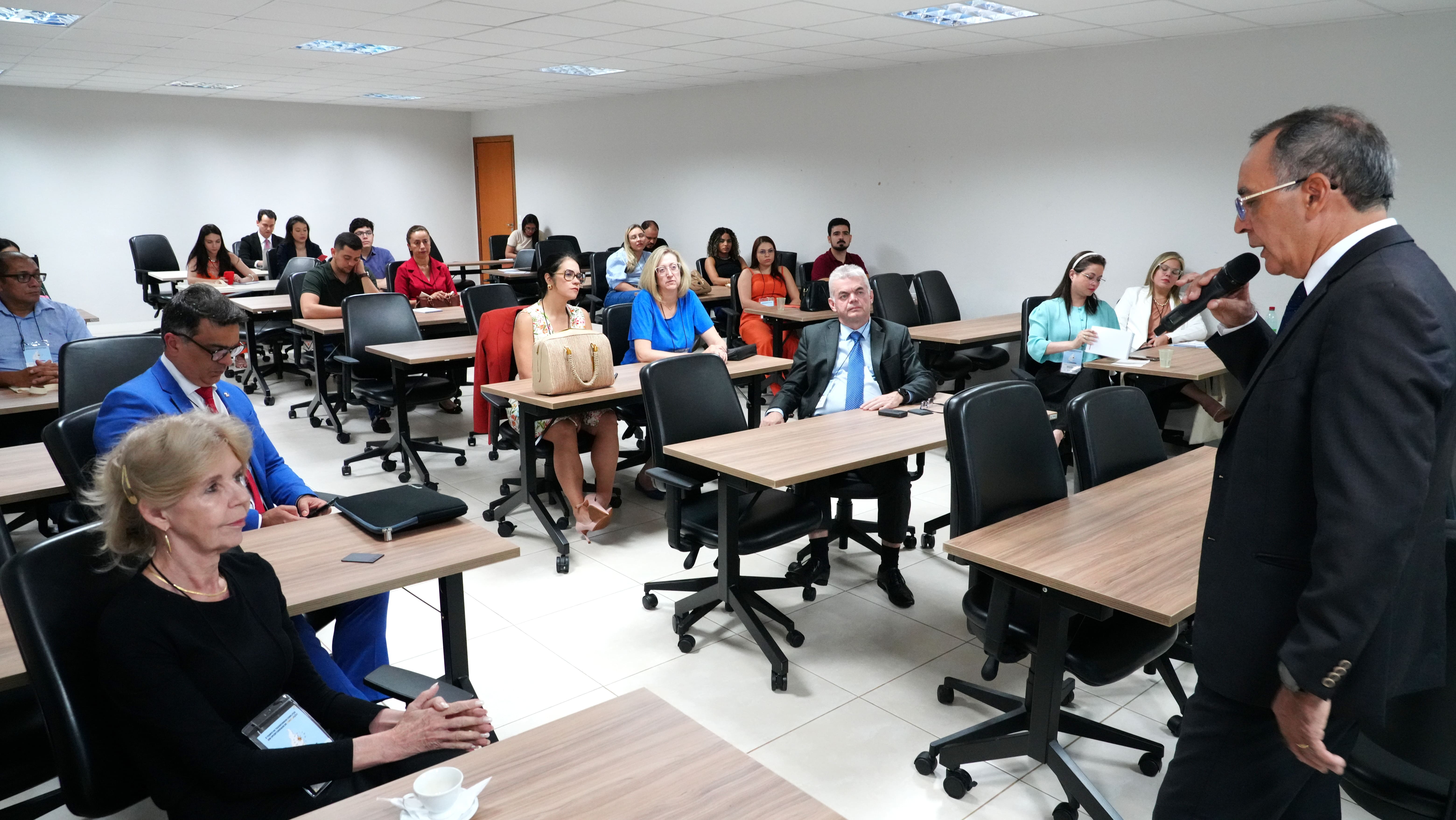 A imagem mostra uma oficina do Fórum Nacional de Mediação e Conciliação realizada em uma sala de aula da Escola Superior da Magistratura Tocantinense. Um homem está em pé, segurando um microfone e falando para os participantes. Várias pessoas estão sentadas em cadeiras, acompanhando a apresentação.
