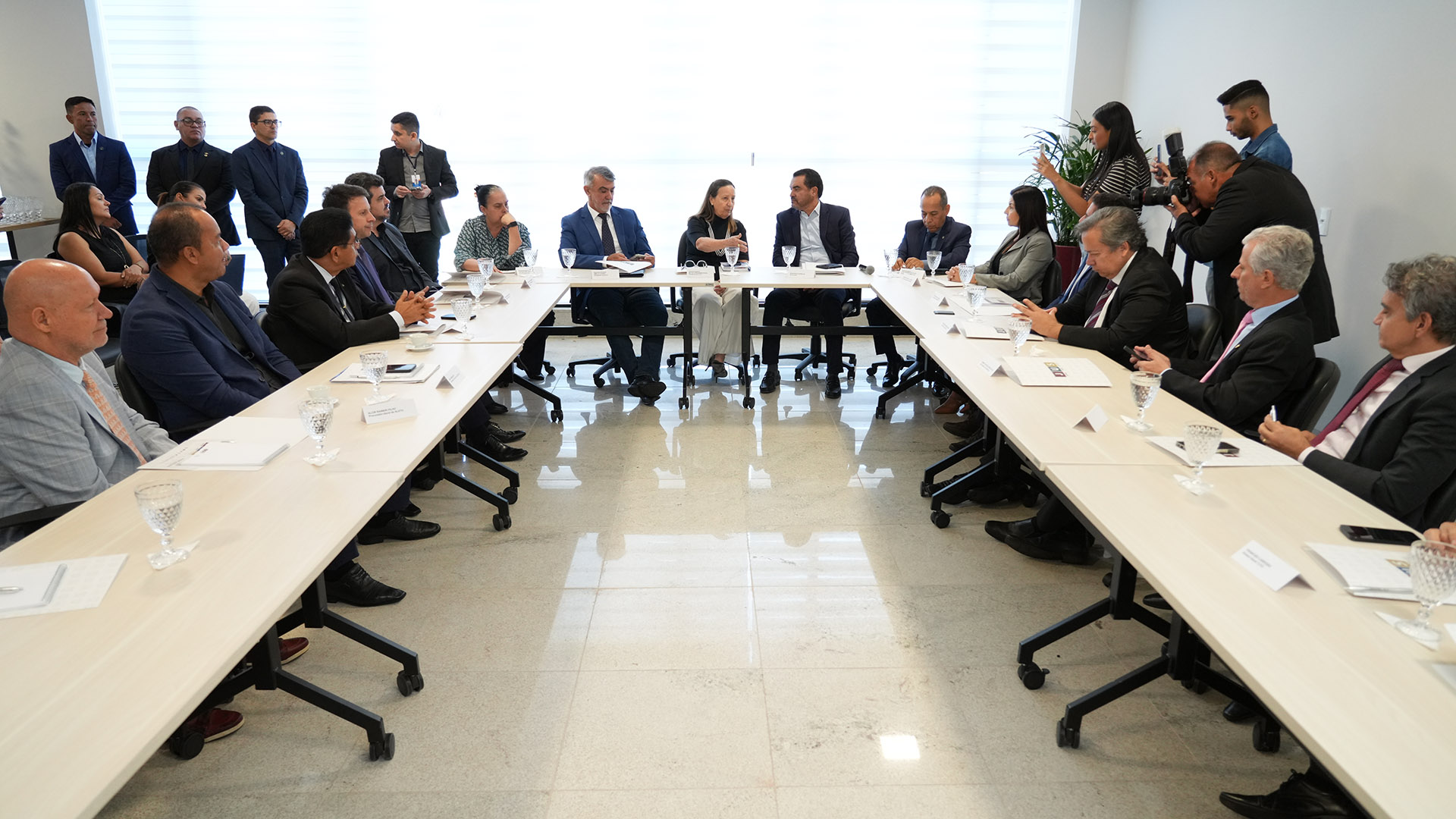 A imagem mostra vários pessoas sentadas em torno de uma mesa retangular em uma sala de reunião.