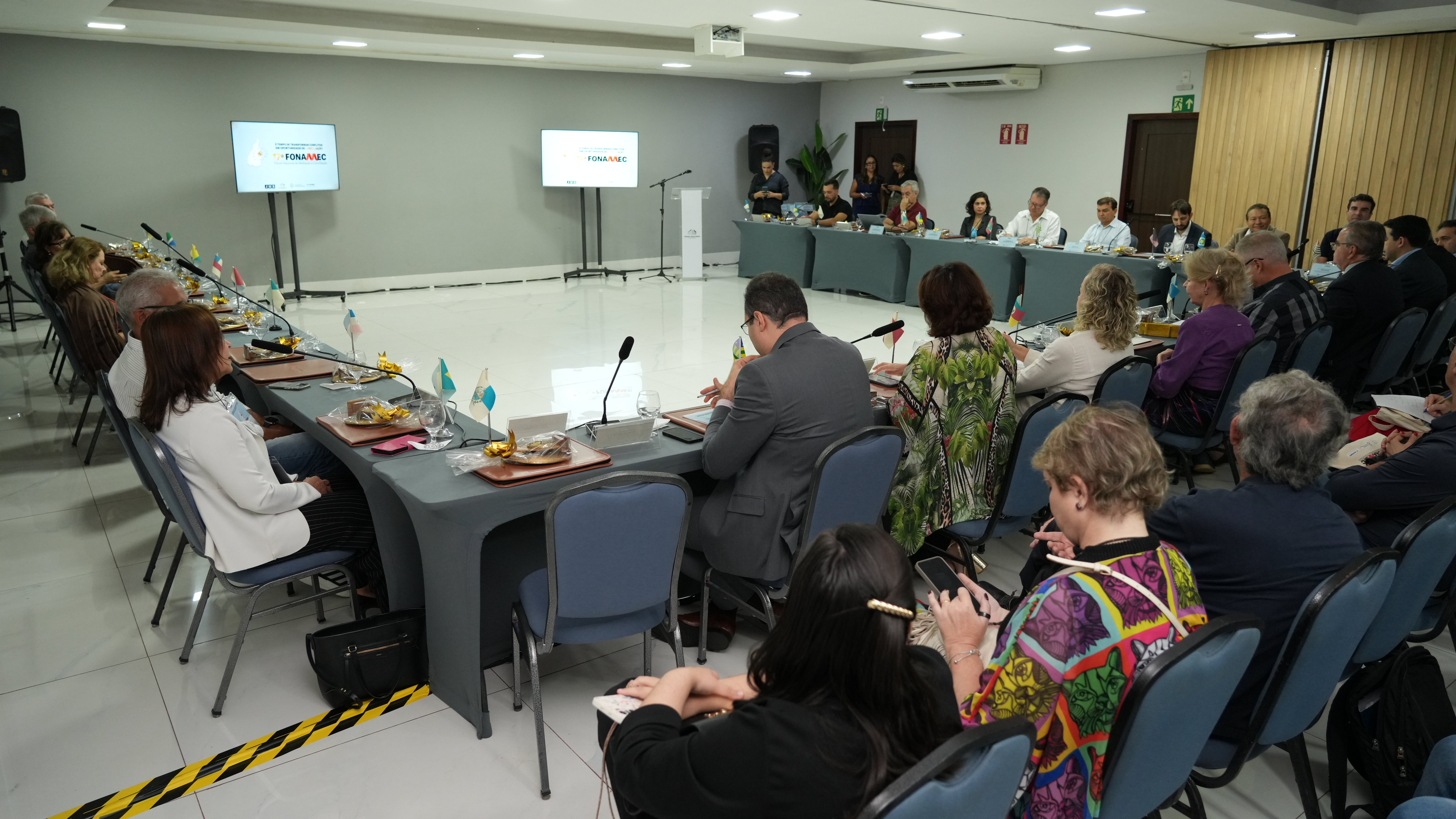 Homens e mulheres sentados de costas em mesa em formato U, em sala de paredes cinzas durante reunião sobre conciliação. Ao fundo, dois telões exibem informações sobre tema da reunião