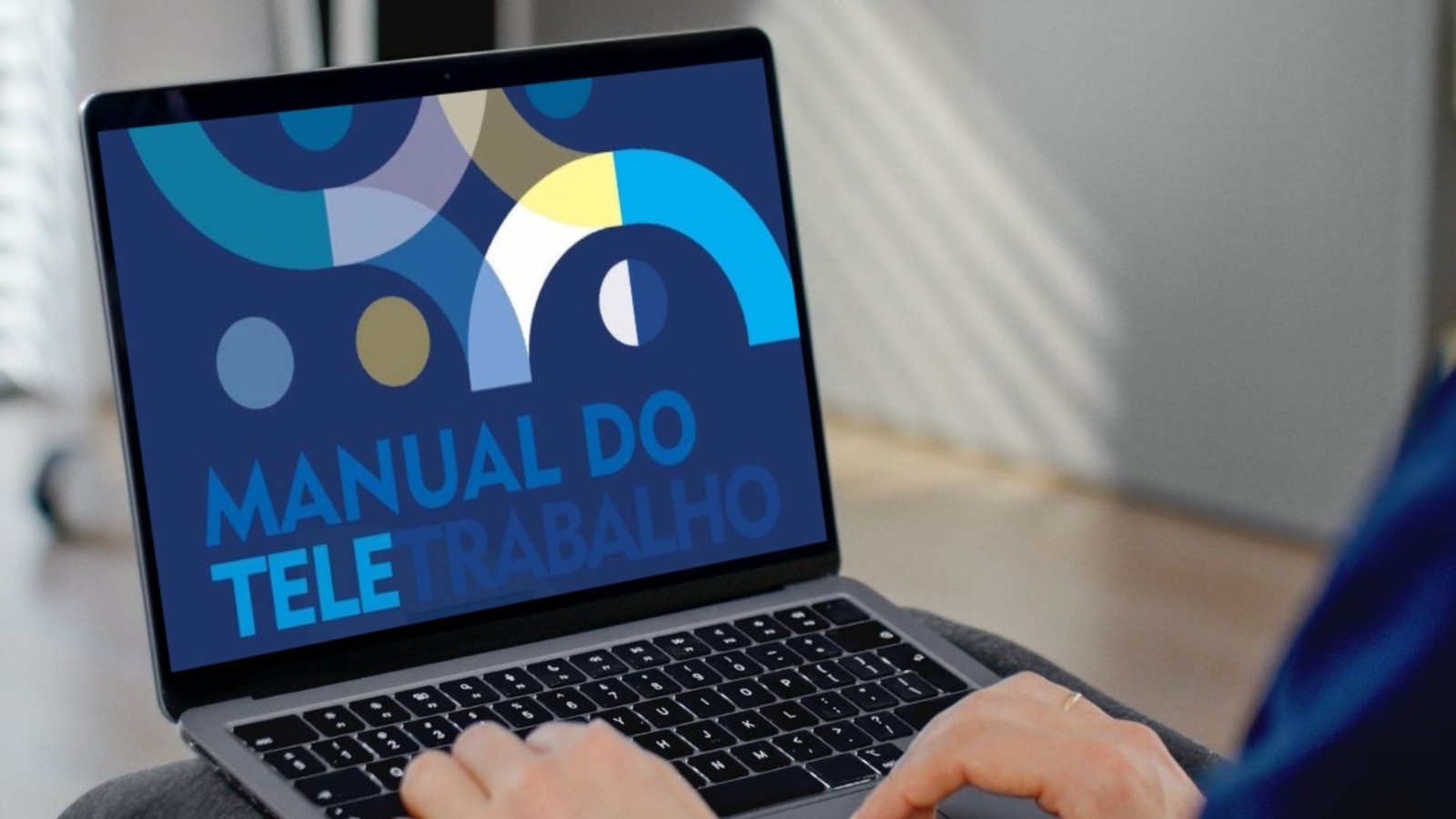 Na imagem aparece uma tela de computador, onde está escrito manual do teletrabalho. Na imagem parece também um teclado e mãos sobre ele.