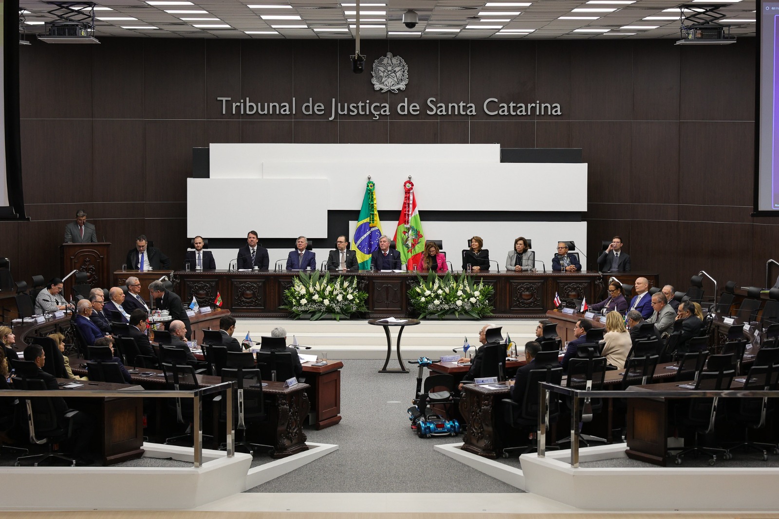 Imagem horizontal de uma sessão solene realizada no plenário do Tribunal de Justiça de Santa Catarina. No centro do plenário, há uma mesa elevada com nove autoridades sentadas, ao fundo das bandeiras do Brasil e de Santa Catarina. A parede é de cor escura, com o brasão da República e a inscrição “Tribunal de Justiça de Santa Catarina” em destaque. À frente, em semicírculo, estão dispostas várias mesas com demais participantes sentados, incluindo magistrados, servidores e convidados. O ambiente é amplo, iluminado e formal, com arranjos florais na mesa principal e decoração sóbria, reforçando o caráter oficial do evento.