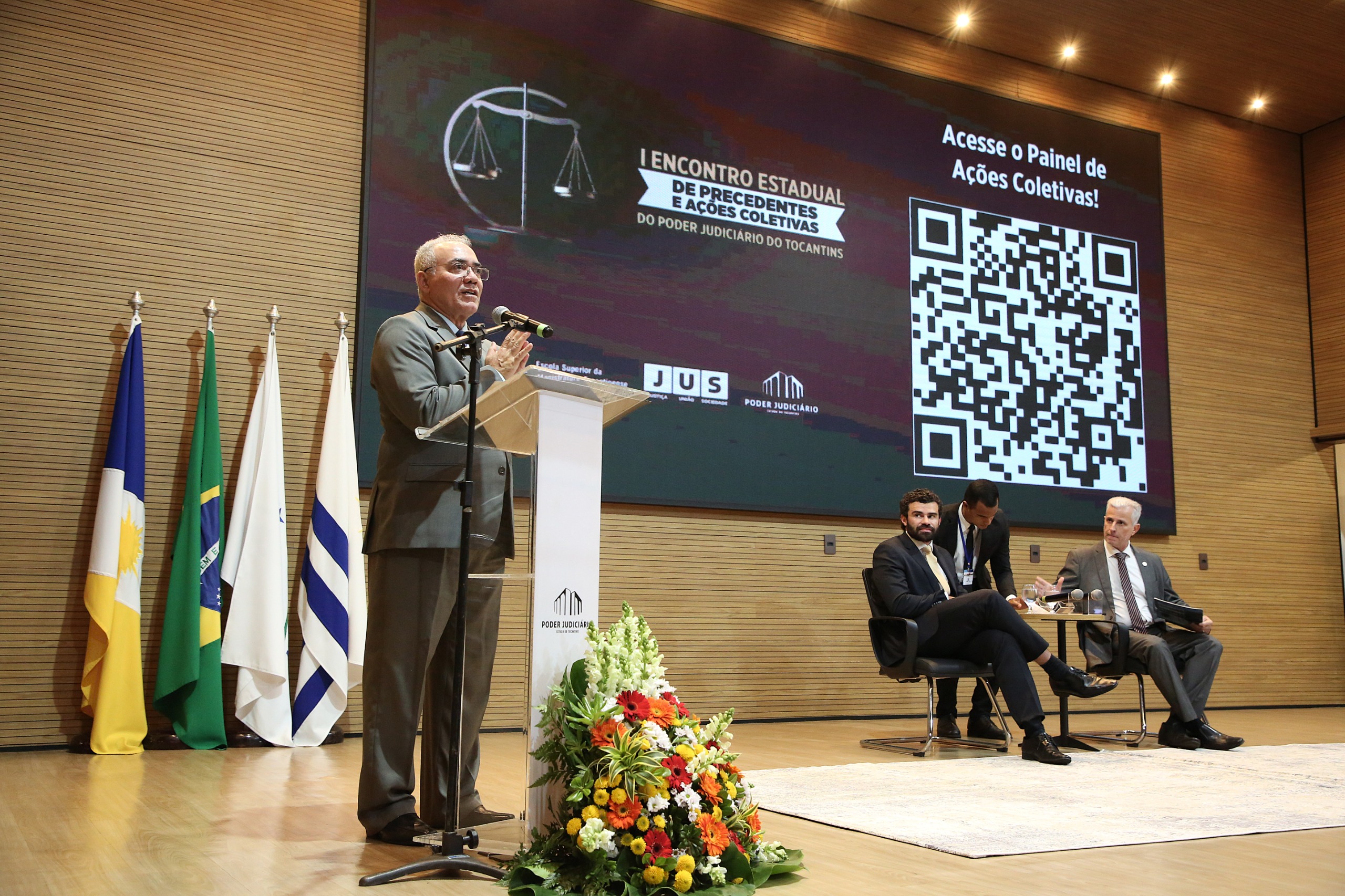 na imagem o desembargador Adolfo Amaro Mendes discursa no palco durante o I Encontro Estadual de Precedentes e Ações Coletivas. Ao fundo, um telão exibe o nome do evento e um QR Code para acesso ao painel. No canto, três participantes acompanham a apresentação. Um arranjo de flores decora a frente do púlpito.