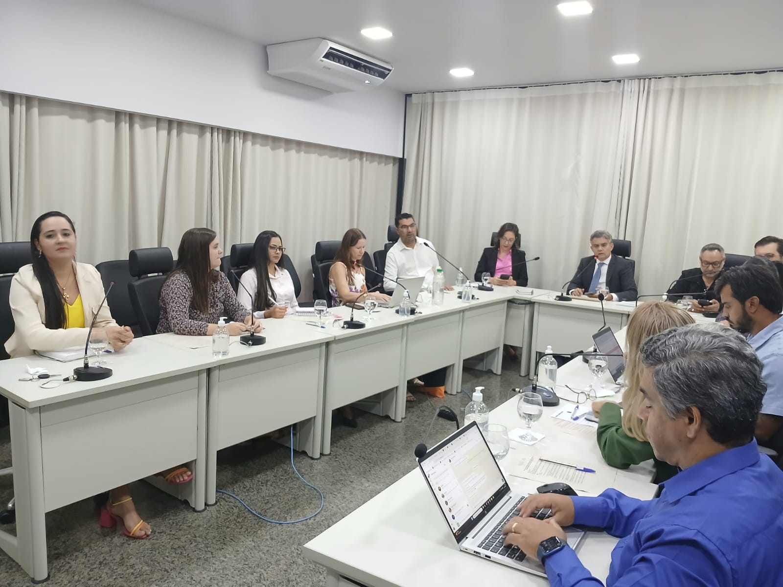 Homens e mulheres sentados em mesa branca em formato U, em sala de paredes brancas e persiana bege