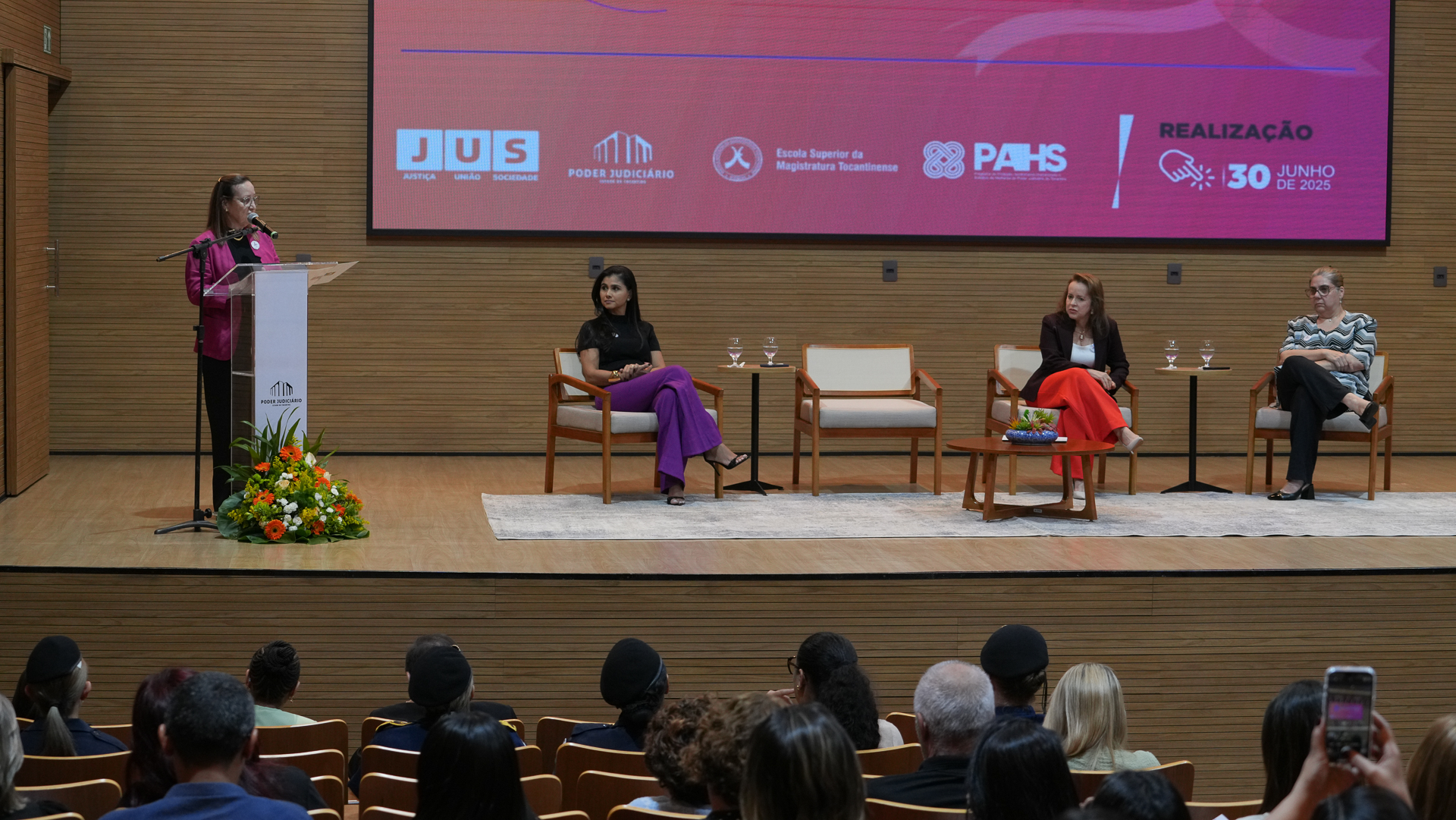 A imagem mostra a abertura do evento Fortalecendo Laços, no auditório do Tribunal de Justiça do Tocantins. No palco, três mulheres estão sentadas em cadeiras e uma quarta ocupa o púlpito, conduzindo a fala. À frente do palco, o público acompanha atentamente a cerimônia, com várias pessoas distribuídas nas cadeiras do auditório.