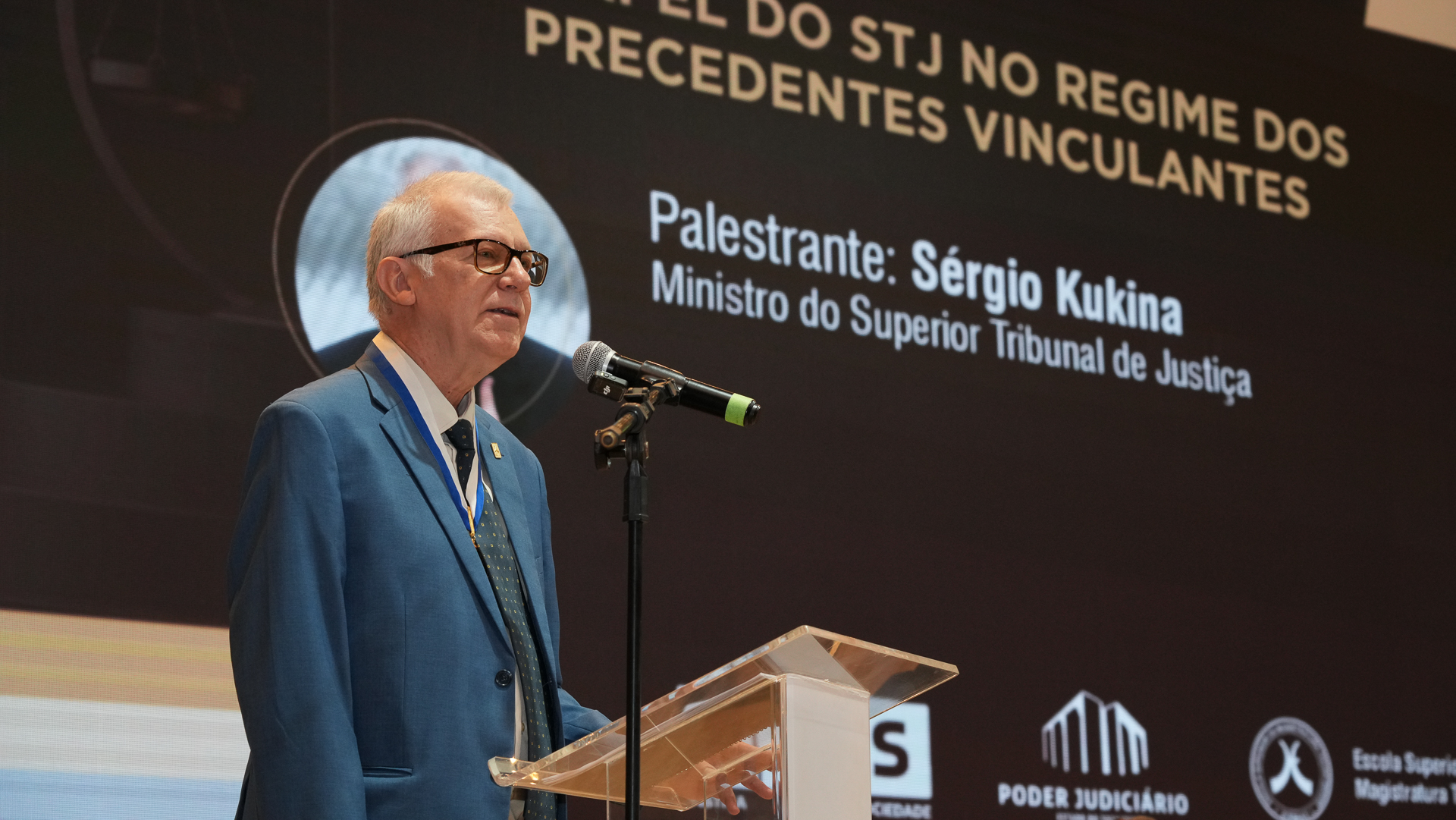 Imagem do ministro Sérgio Kukina falando ao microfone; ele é um home de pele clara, cabelos brancos, usa óculos e terno azul; ao fundo um banner do evento