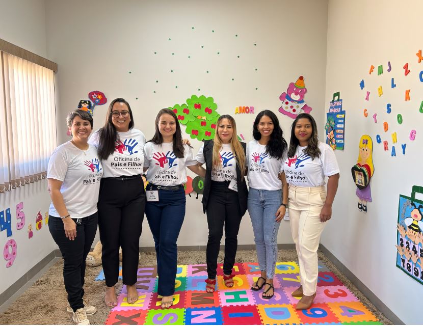 Seis mulheres estão em pé, lado a lado, sorrindo para a foto em uma sala decorada com elementos infantis. Todas vestem camisetas brancas com a estampa “Oficina de Pais e Filhos”, que traz o desenho de duas mãos coloridas (uma azul e uma vermelha). Algumas usam calças jeans e outras calças sociais, e duas delas também vestem blazer. O chão é coberto por tapetes coloridos de EVA com letras e números, e as paredes estão enfeitadas com figuras de animais, árvore com corações e desenhos infantis. A palavra “AMOR” está em destaque na parede ao fundo. A iluminação natural entra por uma janela com persiana à esquerda.