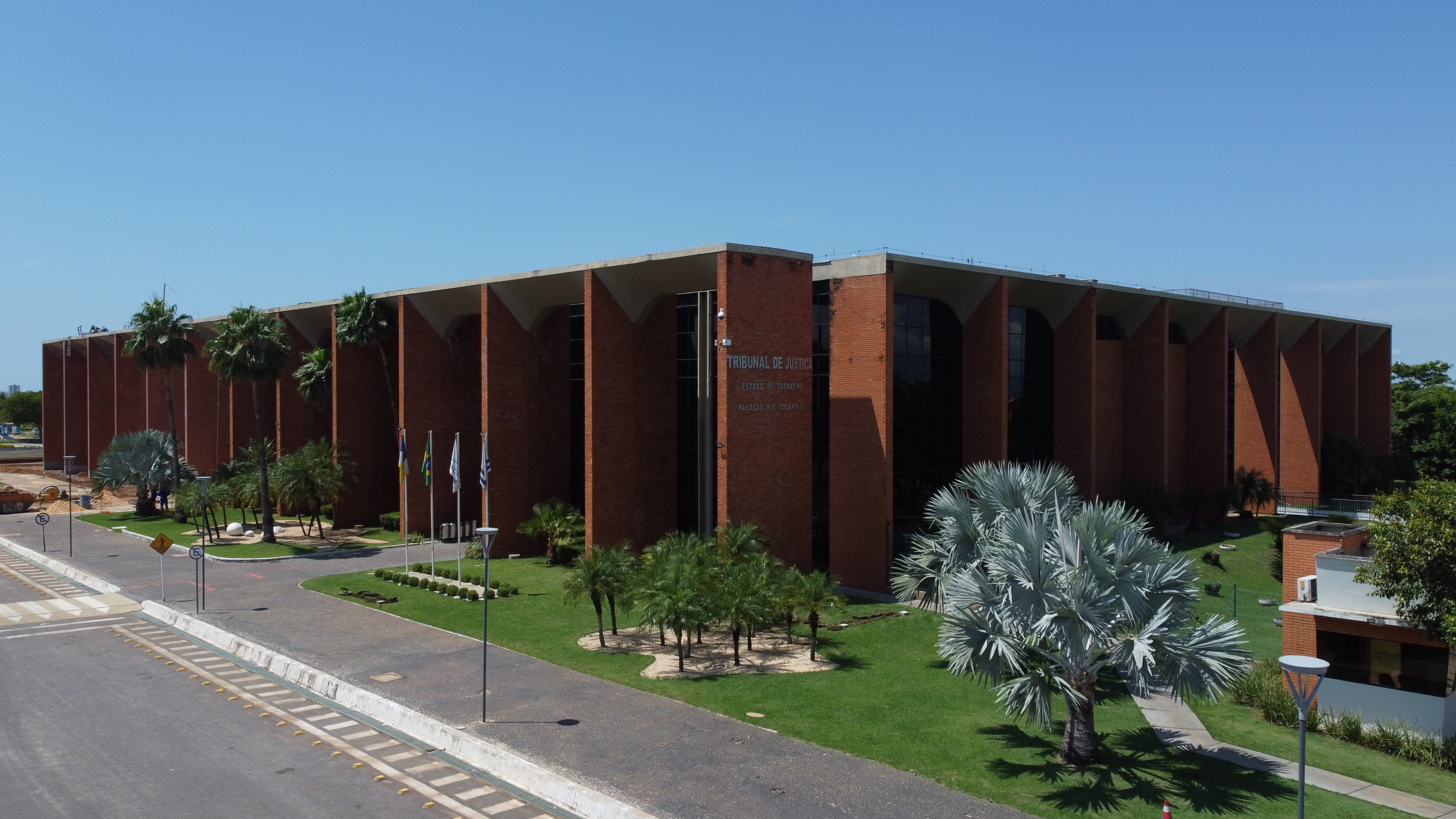 Imagem externa do prédio do Tribunal de Justiça do Tocantins, em Palmas. A construção tem fachada de tijolos vermelhos, colunas altas e jardins com palmeiras e árvores ornamentais. Em frente, estão bandeiras hasteadas e uma via asfaltada. O céu está limpo e ensolarado.