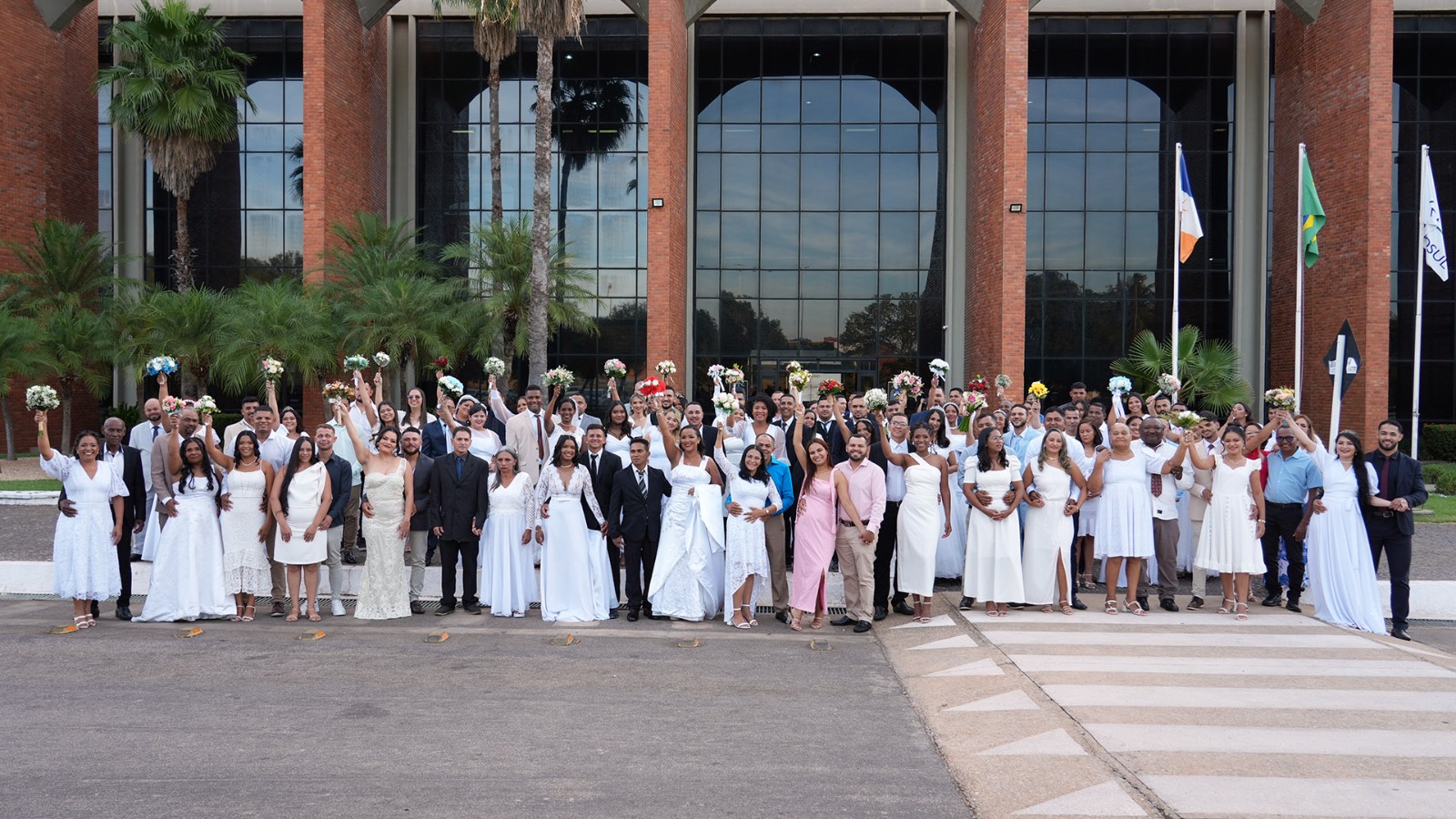 A fotografia mostra dezenas de casais recém-casados, vestidos de branco, posando sorridentes em frente à fachada moderna do prédio do Tribunal de Justiça do Tocantins, em Palmas. O grupo ocupa quase toda a largura da imagem, em clima de celebração e alegria. Muitas noivas seguram buquês erguidos, enquanto noivos e familiares sorriem e se abraçam. Ao fundo, o prédio de tijolos aparentes com amplas janelas de vidro reflete o céu azul. Três mastros com bandeiras e palmeiras completam o cenário festivo do casamento comunitário.