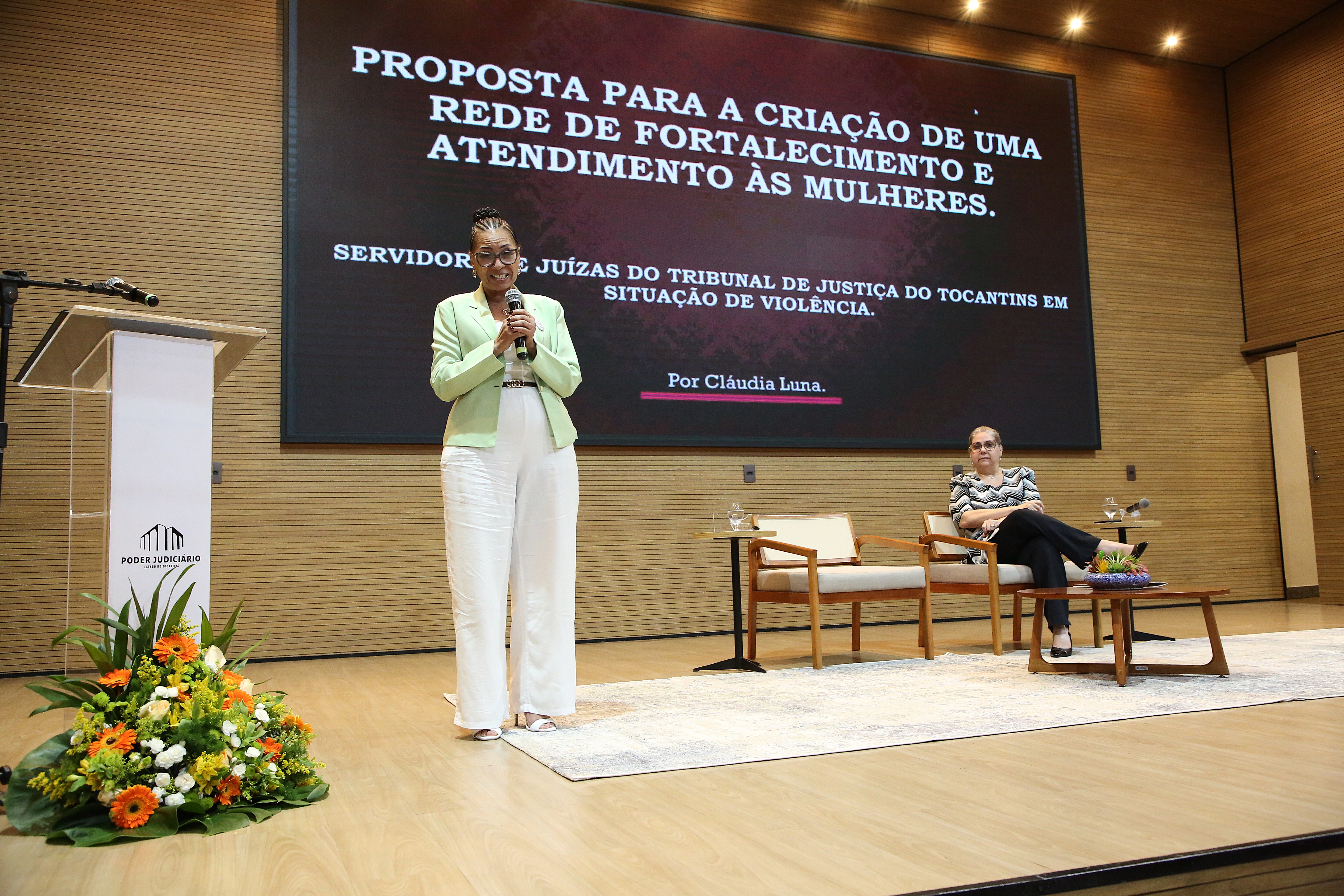 A imagem mostra um momento do evento Fortalecendo Laços, realizado no Tribunal de Justiça do Tocantins. Em destaque, uma mulher está em pé no centro do palco, trajando uma blusa verde-clara e calça branca, enquanto se dirige ao público. Ao fundo, um projetor exibe uma apresentação sobre a criação de uma rede de apoio para mulheres, reforçando o tema central do encontro. Sentada em uma cadeira, também no palco, outra mulher acompanha atentamente a apresentação.