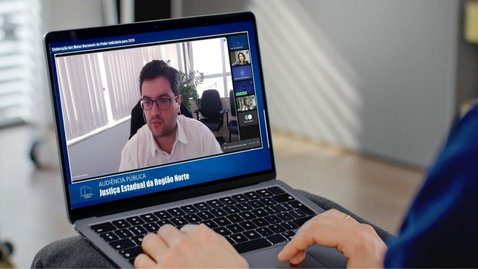 Coordenador do TJ, Renato Alves Gomes, utilizando um notebook no colo participa de uma videoconferência. Na tela do computador, um homem de óculos e camisa branca fala durante uma audiência pública virtual. O fundo da videoconferência mostra uma sala com cadeiras e persianas brancas. A tela exibe a inscrição: "Elaboração das Metas Nacionais do Poder Judiciário para 2026 - Audiência Pública - Justiça Estadual da Região Norte", com o logotipo do Tribunal de Justiça no canto inferior esquerdo.
