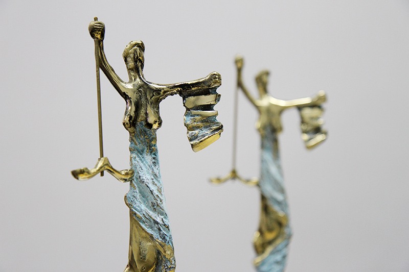 Escultura estilizada da deusa da justiça, representada com olhos vendados, segurando uma balança em uma mão e uma pilha de livros ou processos na outra. A obra é feita em tom metálico dourado com detalhes azul-esverdeados, e está desfocada ao fundo uma segunda escultura semelhante.
