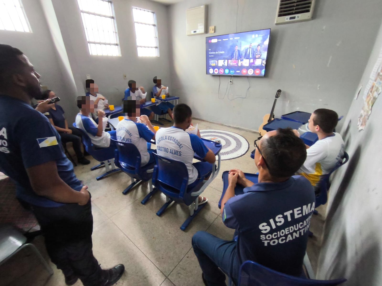 Meninos adolescentes sentados de costas em cadeira azul, em frente a uma TV. Eles estão acolhidos em instituição socioeducativa por terem cometido ato infracional