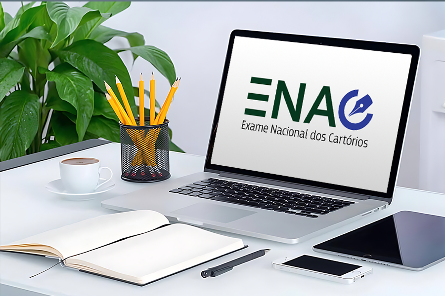 Na imagem aparece uma mesa de trabalho com notebook exibindo a logo do ENAC – Exame Nacional dos Cartórios. Ao lado, caderno aberto, lápis, caneta, xícara de café, celular e tablet. Ao fundo, uma planta verde decora o ambiente.