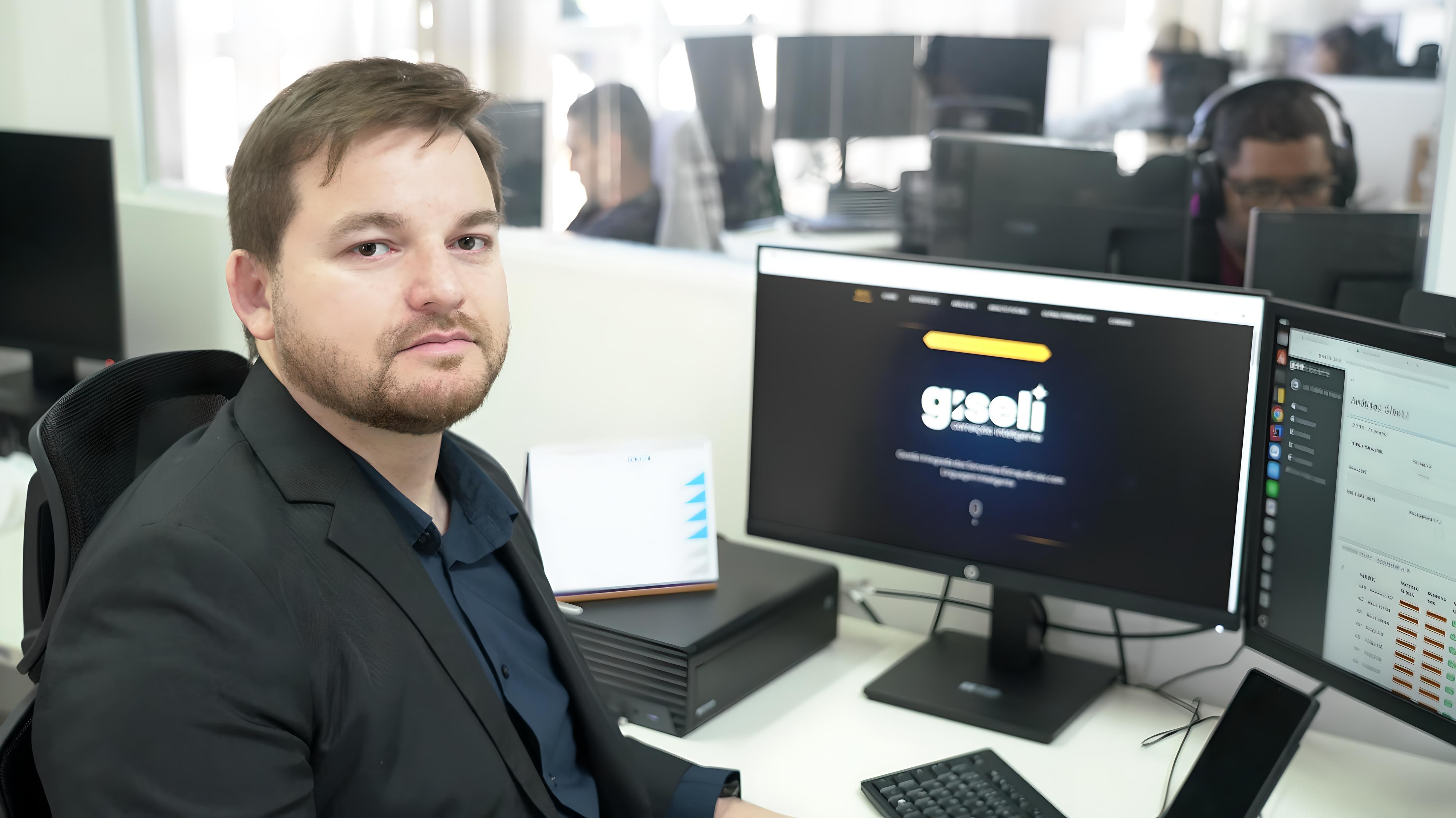 Homem branco de cabelos lisos e castanhos sentado à estação de trabalho, posa para foto diante de computador de duas telas 