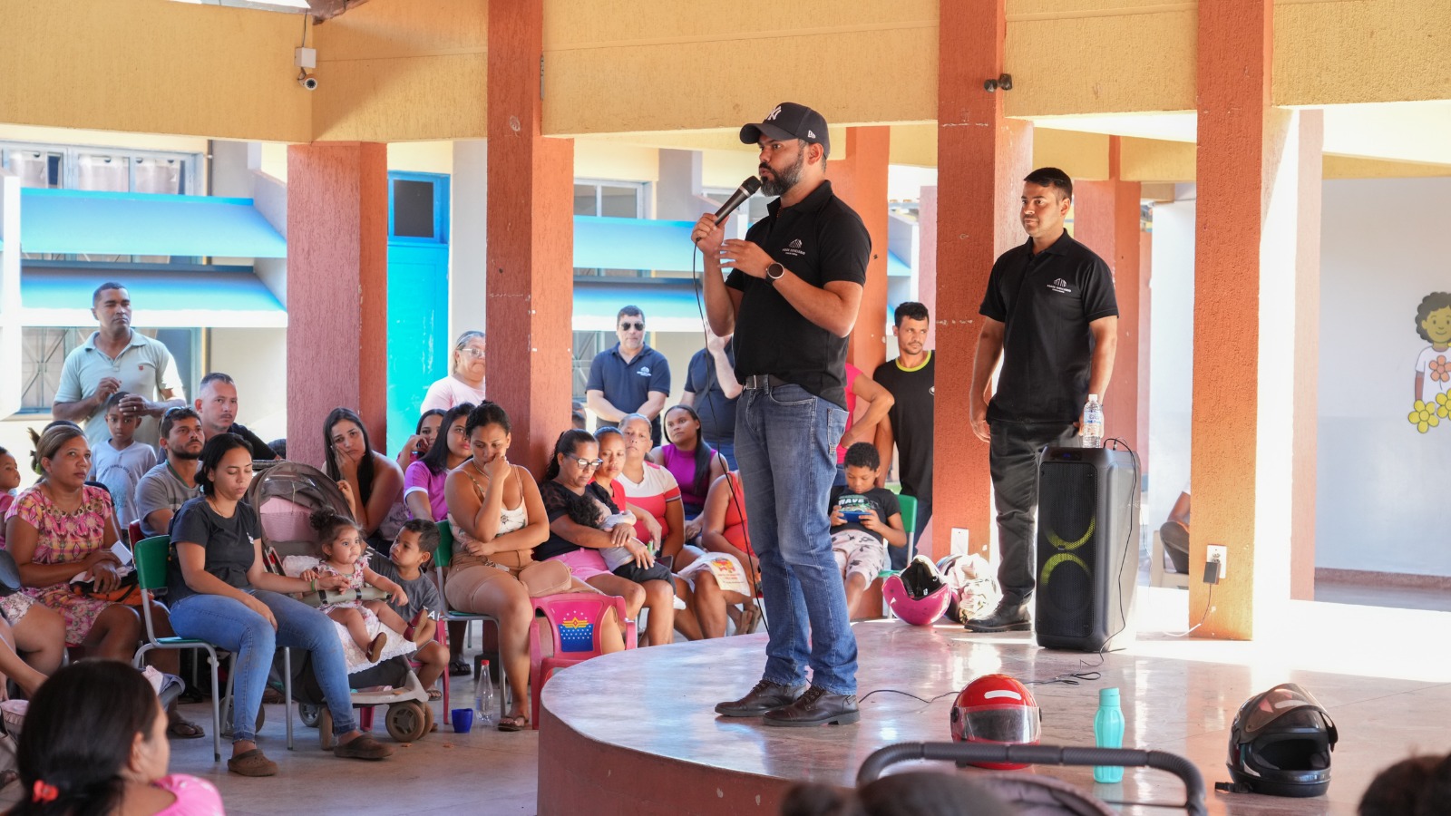 A imagem mostra um homem em pé, sobre um pequeno palco circular, segurando um microfone e falando para um grupo de pessoas. Ele usa boné, camiseta preta e jeans. Ao lado dele, outro homem também de camiseta preta está em pé ao lado de uma caixa de som. O público, composto por mulheres, crianças e alguns homens, está sentado em cadeiras plásticas em volta do palco, em um espaço coberto com colunas alaranjadas. Muitas das mulheres têm crianças no colo ou ao lado, e há carrinhos de bebê e capacetes no chão. O ambiente sugere uma reunião comunitária ou ação social, em local como uma escola ou centro comunitário.