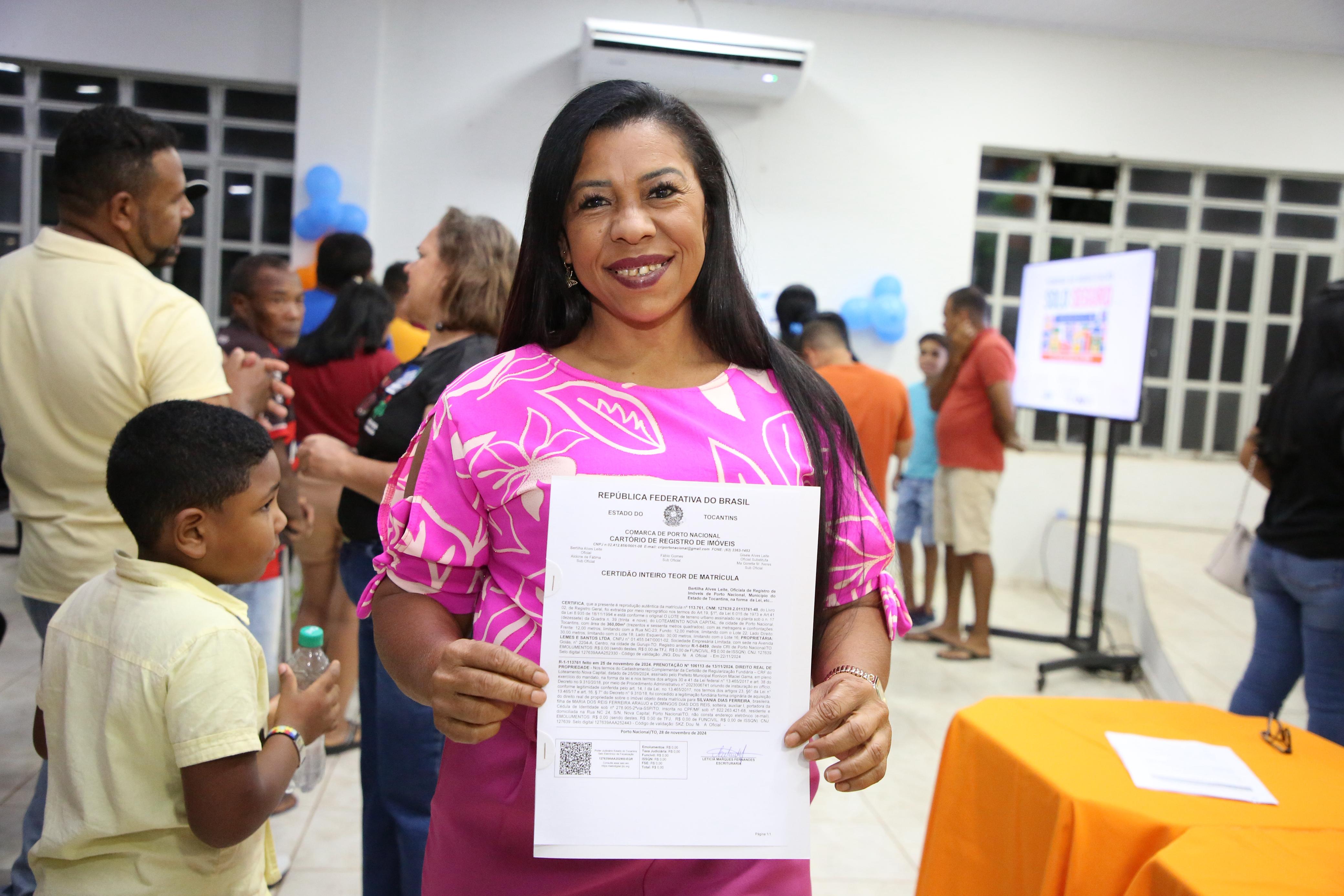 Mulher sorri enquanto segura um documento oficial. Ela veste blusa rosa com estampa clara e está em um ambiente interno com outras pessoas ao fundo, em clima de evento comunitário. Há balões decorativos, mesa com toalha laranja e uma tela digital ao fundo.
