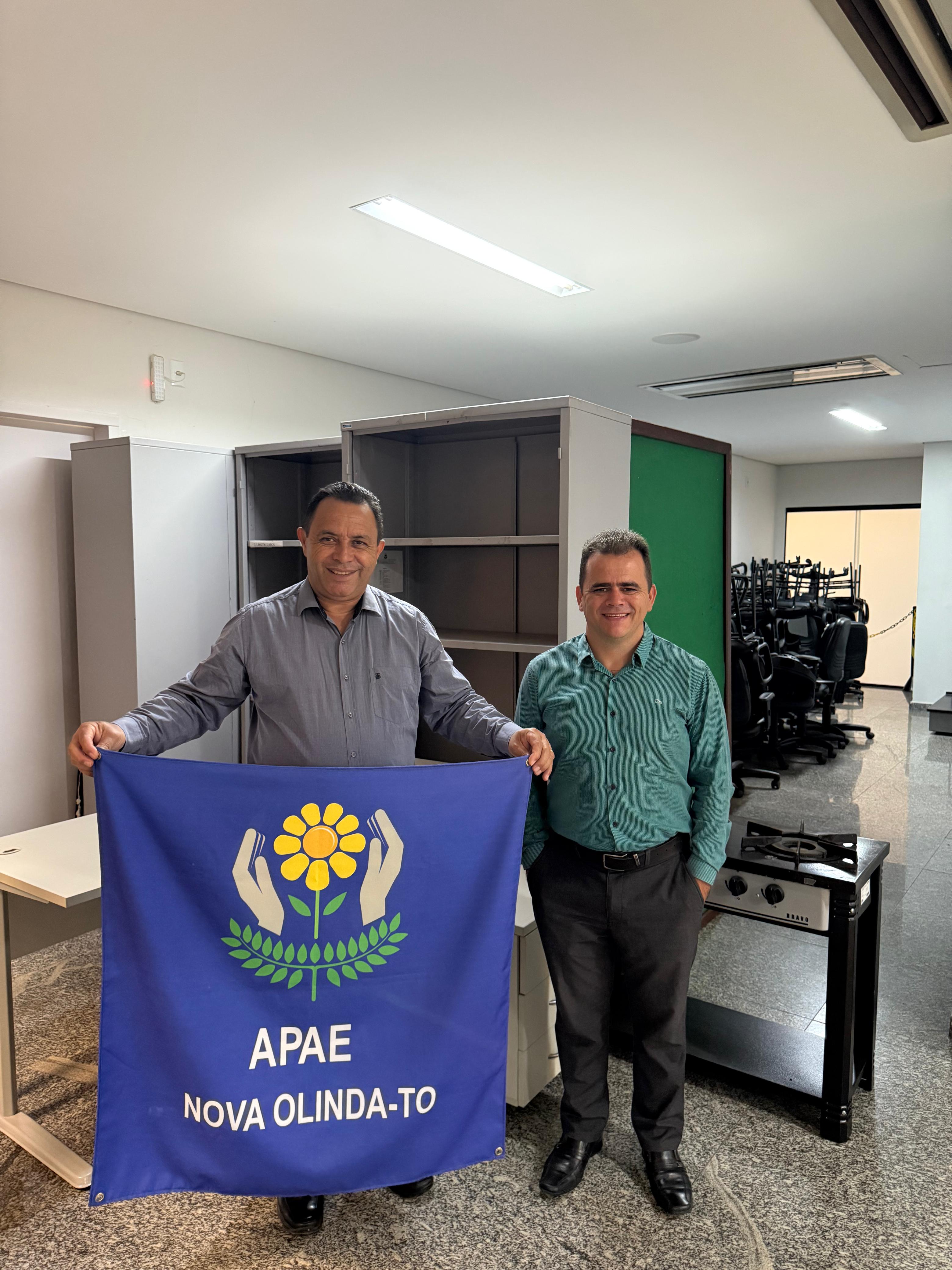 A imagem mostra dois homens estão em pé lado a lado em um ambiente interno, com piso de granito e iluminação fluorescente. Ambos sorriem para a câmera. O homem à esquerda segura um banner azul com o logotipo e o nome da APAE de Nova Olinda-TO. Ao fundo, há móveis e equipamentos como armários, mesas, cadeiras empilhadas e um fogão, indicando um contexto de doação de bens.
