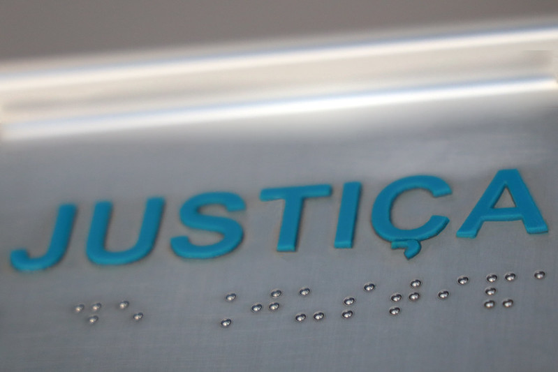 Foto em close de uma placa metálica com a palavra “JUSTIÇA” escrita em letras maiúsculas azuis em relevo. Abaixo da palavra, há a mesma inscrição em braille, com pequenos pontos metálicos em alto-relevo. A imagem transmite a ideia de acessibilidade e inclusão no sistema de Justiça.