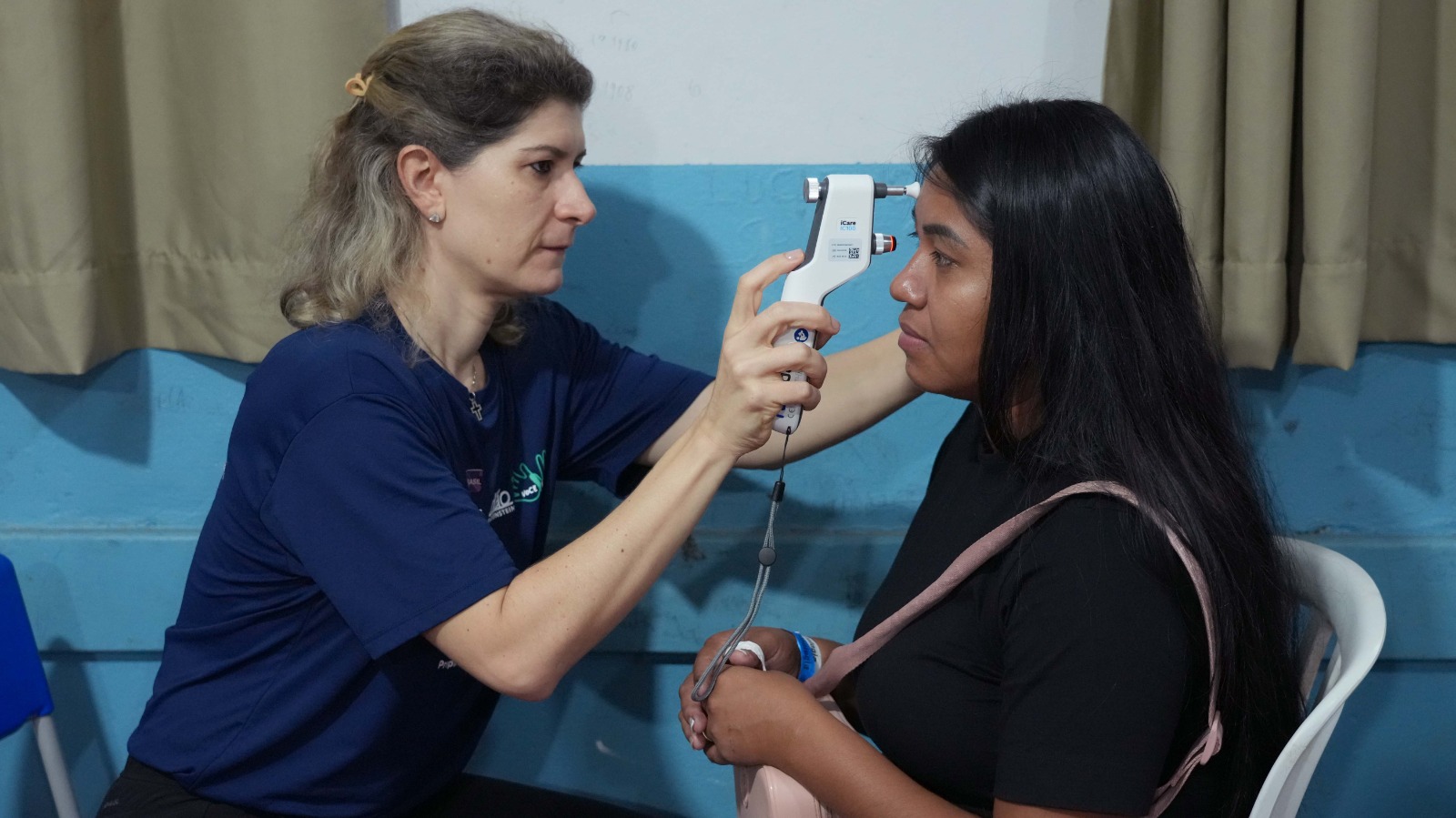 Uma mulher indígena de cabelos longos e escuros está sentada enquanto tem um dos olhos examinado por um profissional de saúde, durante a Semana Nacional da Saúde Indígena