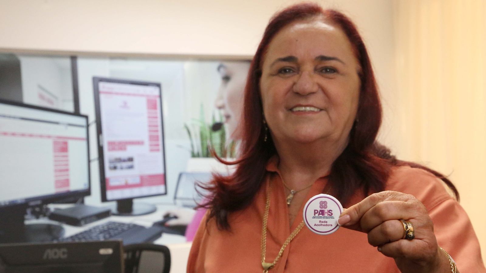 A imagem mostra uma mulher sorrindo enquanto segura o botton de identificação como agente da Rede Acolhedora do Programa de Proteção, Acolhimento Humanizado e Solidário (PAHS).