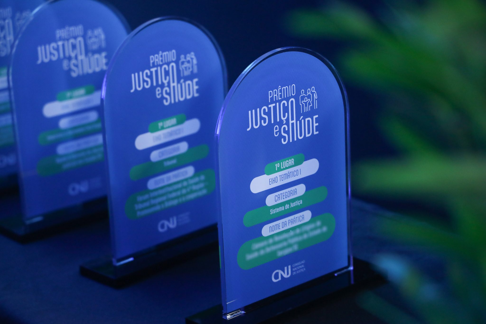 A imagem exibe diversos troféus de acrílico do “Prêmio Justiça e Saúde” do CNJ, organizados em um arranjo. Cada troféu apresenta informações do eixo temático, a categoria e o nome da prática premiada. As cores predominantes são azul, verde e branco.