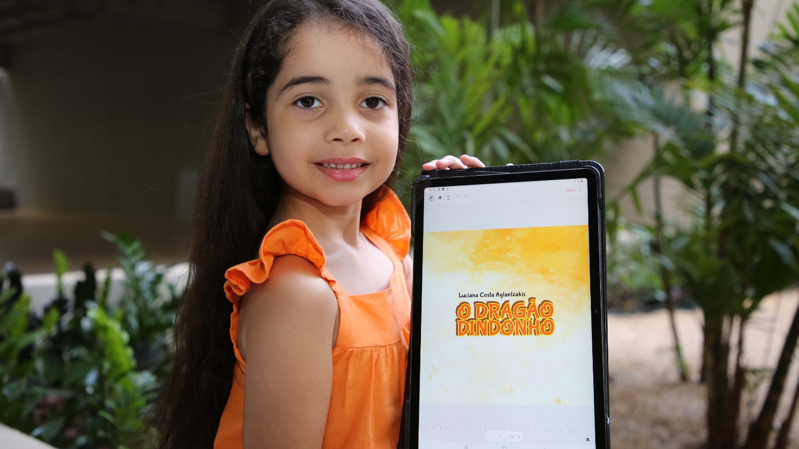 A imagem mostra uma menina sorrindo e segurando um tablet que mostra a imagem da capa do livro infantil ‘O Dragão Dindonho’.