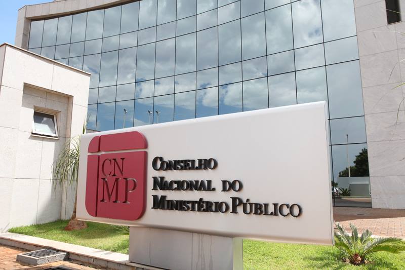 Foto externa da sede do Conselho Nacional do Ministério Público (CNMP), em Brasília. Em primeiro plano, destaca-se uma placa institucional com o logotipo do órgão em vermelho e as palavras “Conselho Nacional do Ministério Público” em letras pretas. Ao fundo, vê-se a fachada espelhada do prédio, refletindo o céu com nuvens. O ambiente é arborizado, com gramado e pequenas plantas ornamentais próximas à entrada.