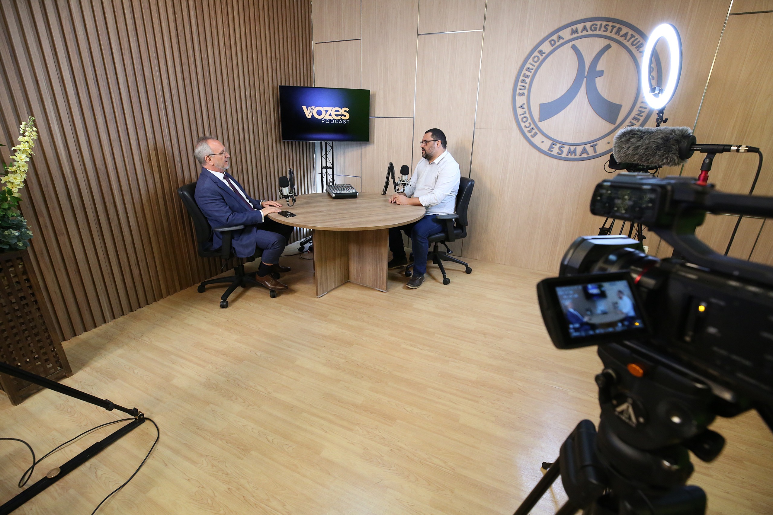 A imagem mostra um estúdio de gravação com dois homens sentados em torno de uma mesa redonda, em frente às câmeras e equipamentos de gravação.