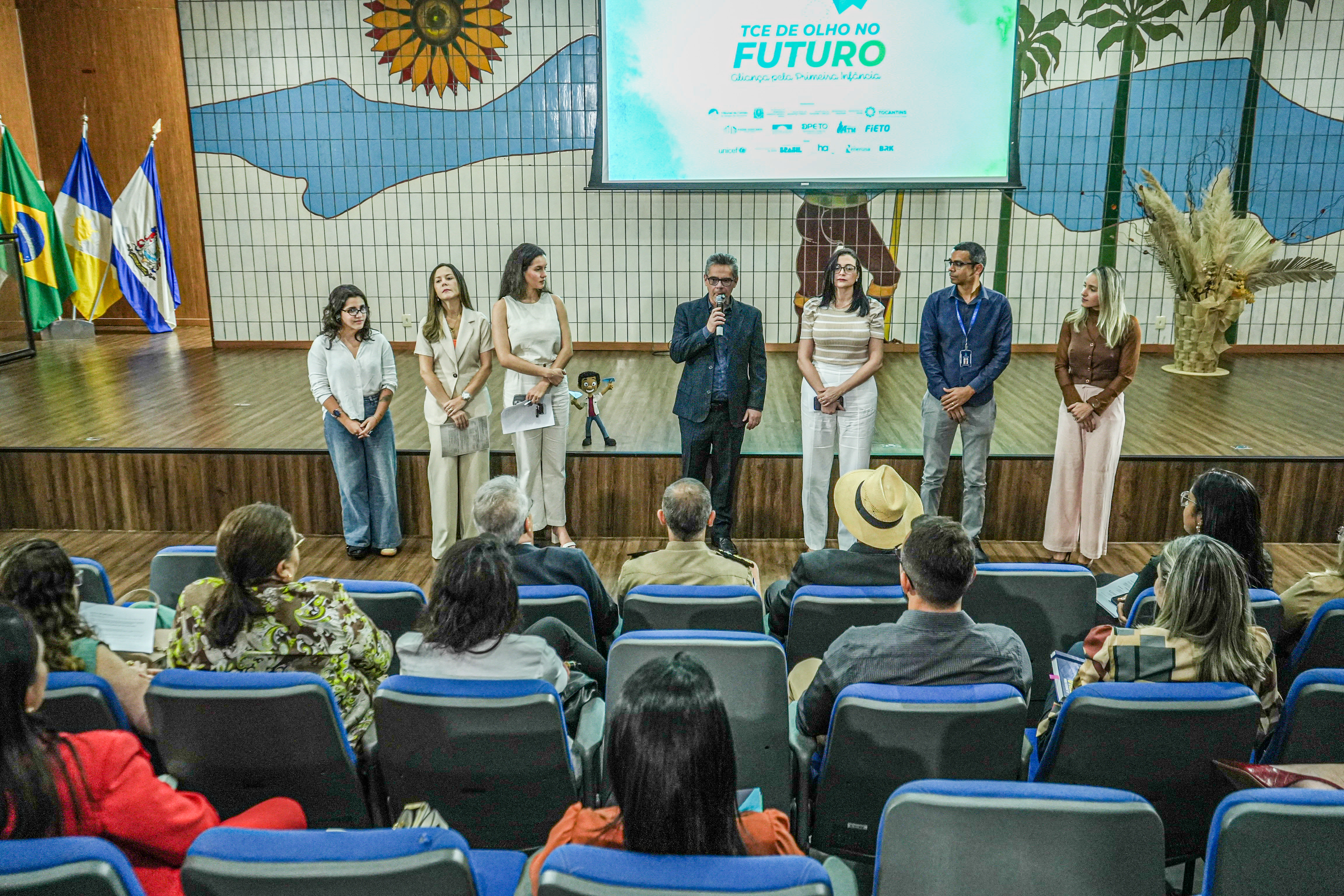 A imagem mostra um auditório durante um evento institucional. No palco, há oito pessoas alinhadas de frente para o público. Uma delas, um homem ao centro, está com microfone em mãos, aparentemente fazendo uma fala. Ao fundo, há um painel com arte colorida de paisagens e um telão que exibe o título "TCE DE OLHO NO FUTURO – Aliança pela Primeira Infância", além de logomarcas de instituições parceiras.