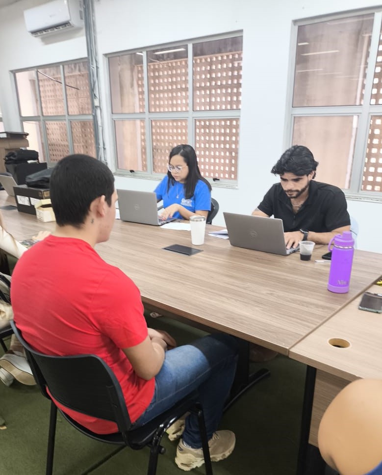 jovem sentado de costa, de camiseta vermelha, fazendo sua matrícula, à frente, do outro lado da mesa, um homem e uma mulher usando computadores