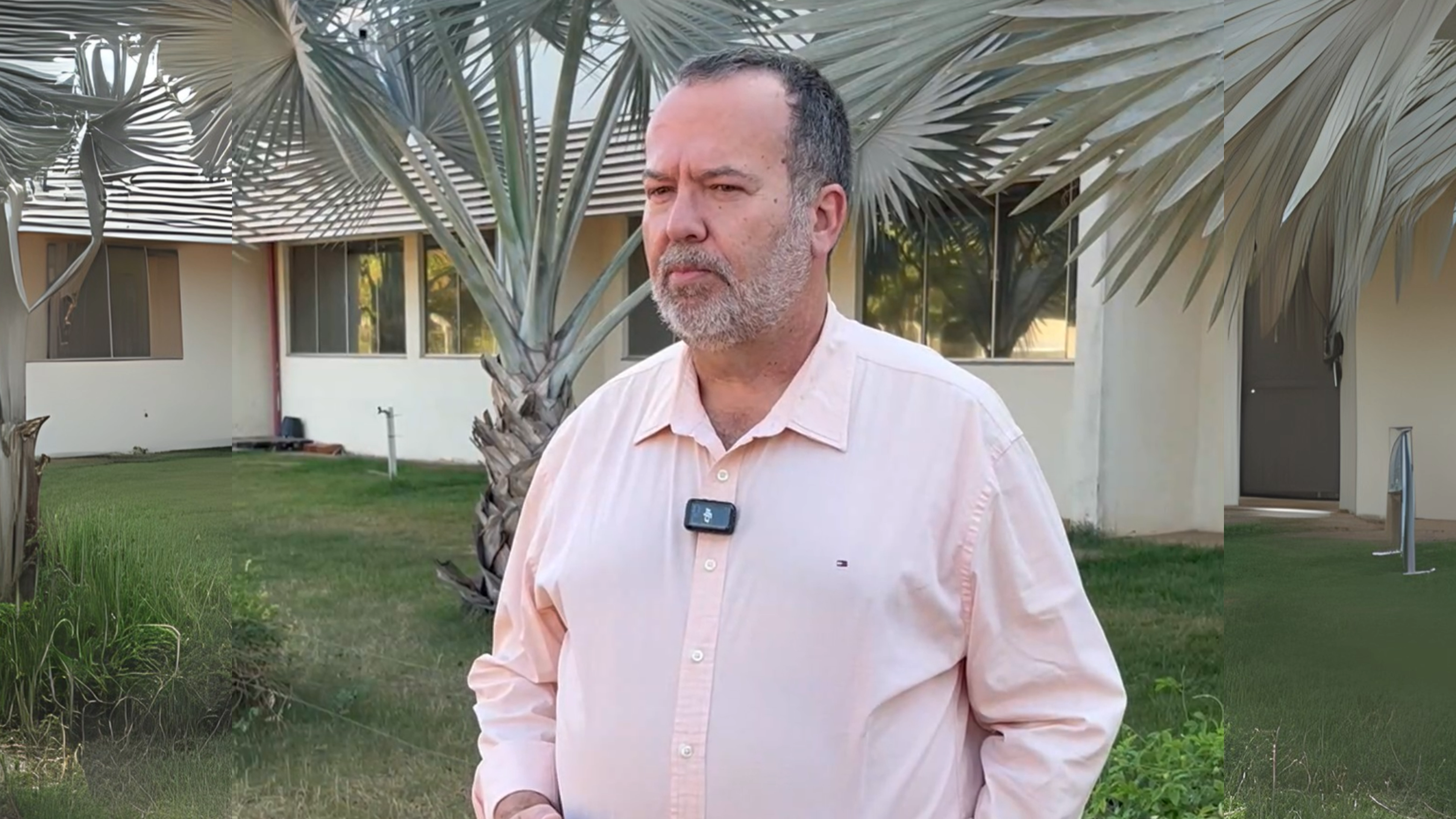 Engenheiro Adrian, homem de pela clara, barba e cabelos grisalhos; ele usa camisa rosa clara e está à frente de um prédio e palmeiras