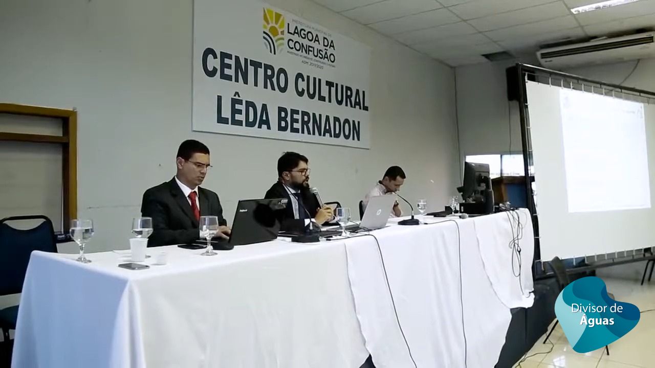 Imagem de uma mesa com a presença de três pessoas sentadas, ao centro o juíz, de terno, fala ao microfone; ao fundo uma placa com o nome Centro Cultural Lêda Bernadon