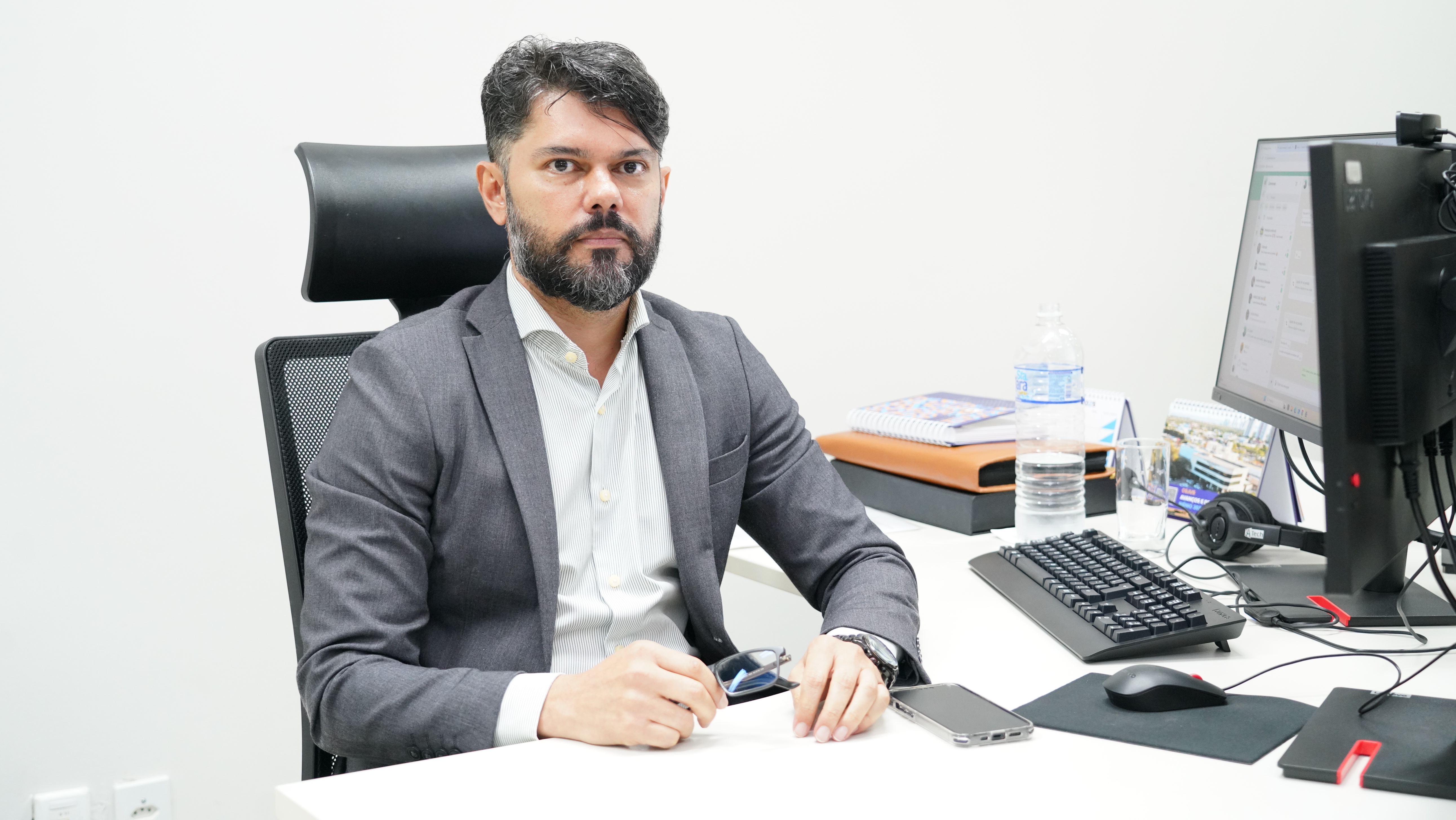 Juiz Wellington Magalhães; homem moreno claro, cabelos e barba escuros; ele está sentado, ao lado de um computador, e usa camisa branca e blazer cinza. 