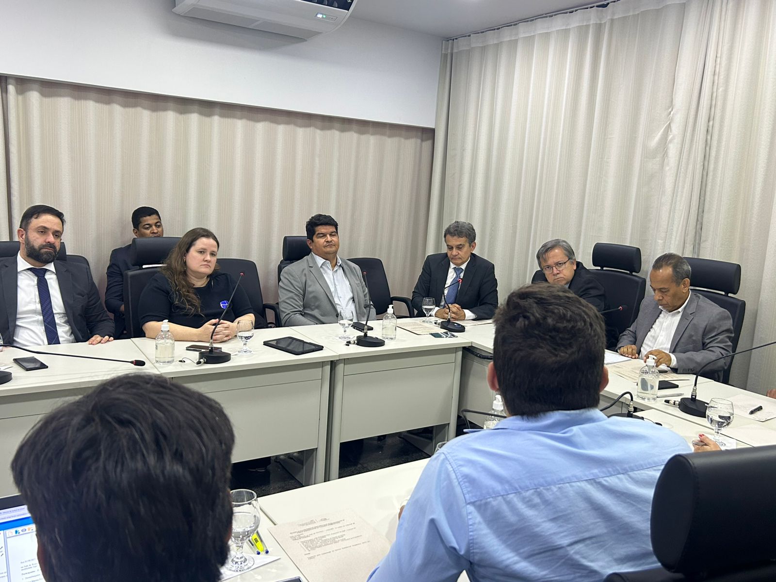 Imagem aberta de uma sala de reuniões onde várias pessoas estão sentadas e conversando; na imagem aparecem mesas brancas, paredes e cortinas brancas