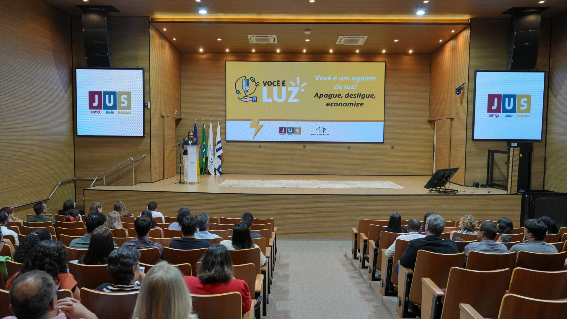 Imagem aberta do auditório do TJTO em que aparece parte do público e ao fundo uma pessoa falando ao microfone, no palco, e monitores projetando imagens