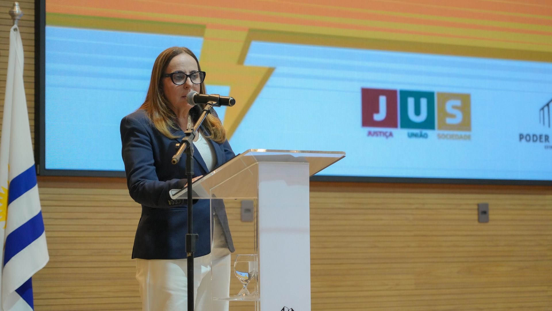 Desembargadora Angela Pudente fala ao microfone; ela usa óculos , roupa branca e terninho azul