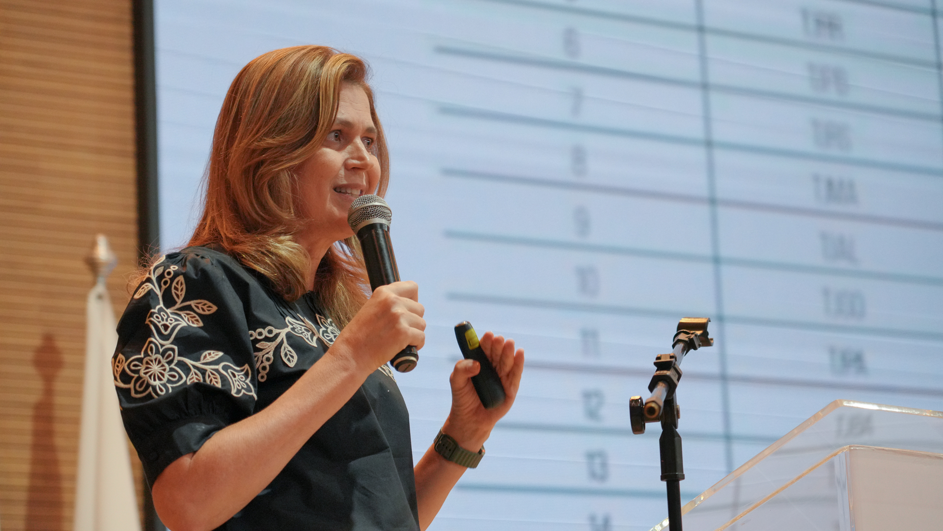 Imagem de uma mulher falando ao microfone; ela tem cabelos e pele clara e usa blusa verde 