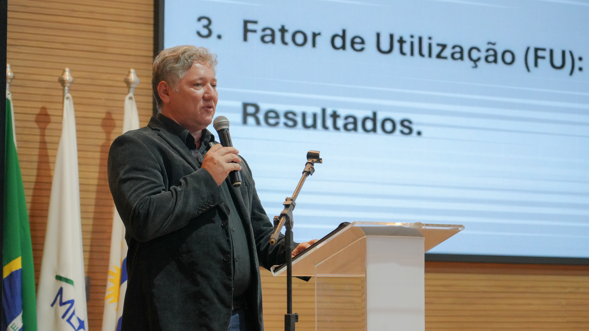 Imagem de um homem de roupa escura, pele clara e cabelos grisalhos, falando ao microfone 