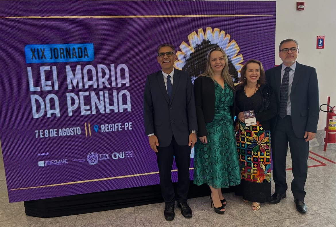 A imagem mostra quatro pessoas sorrindo ao lado de um banner da XIX Jornada Lei Maria da Penha, que acontece nos dias 7 e 8 de agosto, em Recife-PE. O banner traz a imagem de uma mulher negra com flores. Ao lado, há um vaso com plantas. O local tem paredes claras com detalhes amarelos e piso de granito. Ao fundo, outras pessoas circulam pelo ambiente.