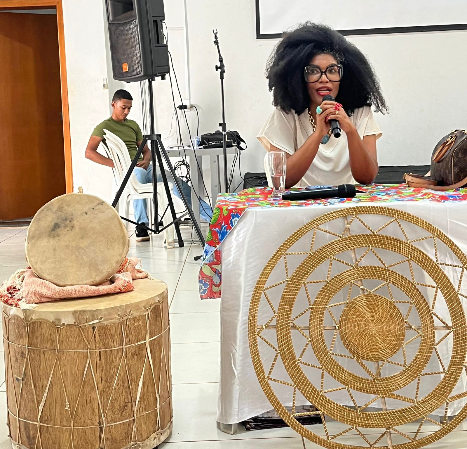 Juiza Renata Nascimento está sentada, falando ao microfone, durante palestra magna no evento