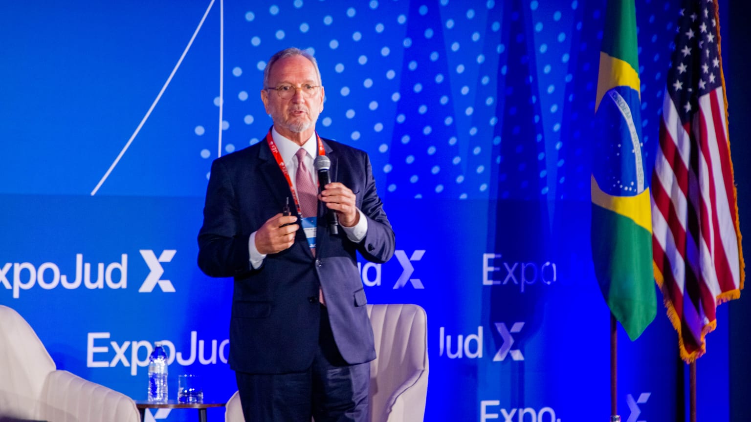 Foto do desembargador Pedro Nelson palestrando em evento. Ele é um homem de pele clara, cabelos e barba grisalhos; ele usa camisa branca, grava em tons de vermelho e terno azul