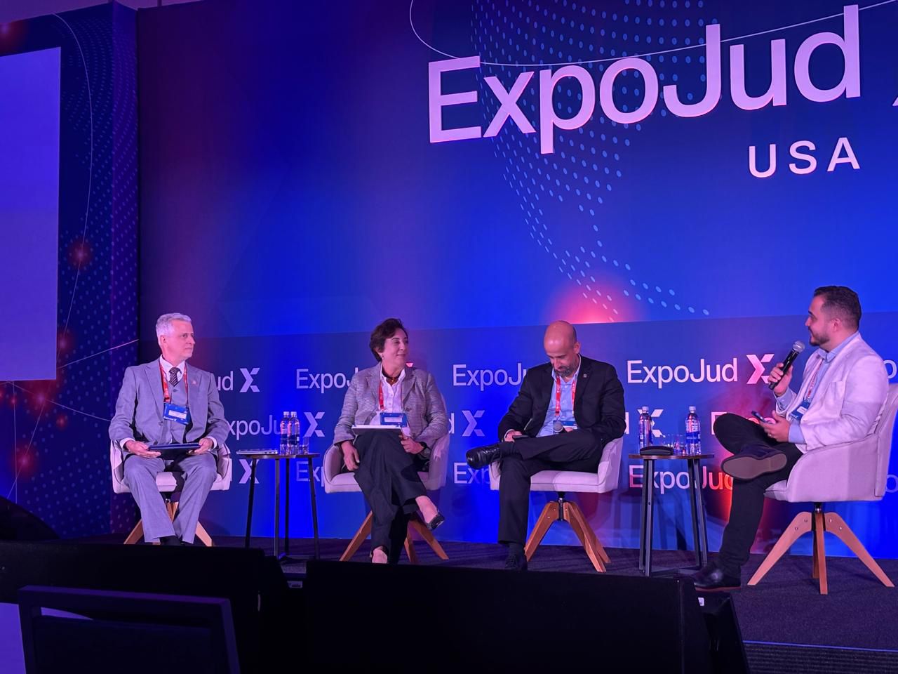 Imagem aberta em que aparecem quatro pessoas, quatro homens e uma mulher. Todos estão sentados participando de um painel em um evento do Judiciári