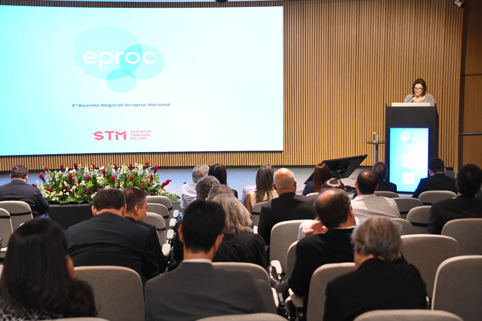 A imagem mostra o auditório do Superior Tribunal Militar, em Brasília, durante a 4ª Reunião Negocial do Sistema e-Proc 2025. Na plateia, dezenas de participantes estão sentados em fileiras, acompanhando a programação. No palco, uma pessoa discursa atrás de um púlpito, e ao fundo, uma grande tela exibe imagens relacionadas ao sistema eproc.