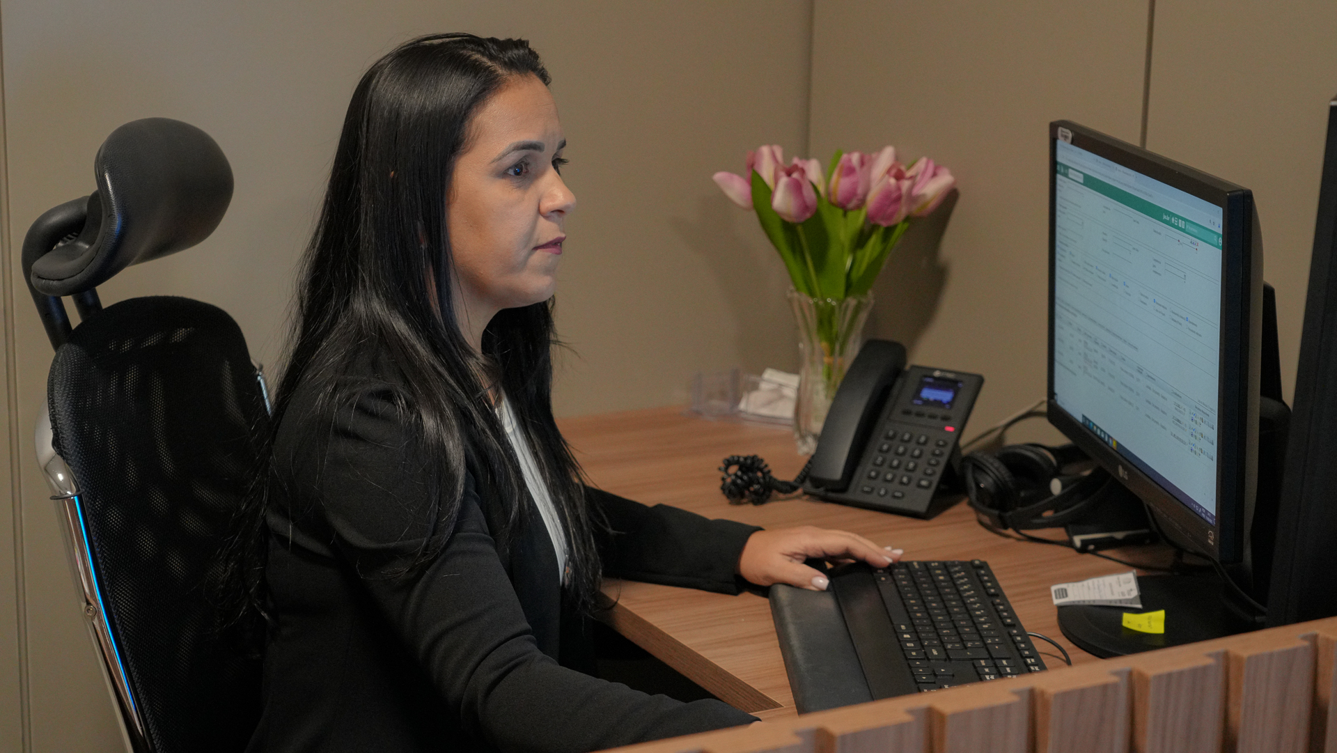A imagem mostra uma mulher de cabelos longos e pretos, vestindo blazer preto, sentada em uma cadeira de escritório com encosto ergonômico. Ela está concentrada olhando para um monitor de computador, digitando no teclado. Sobre a mesa de madeira, há um telefone fixo, um vaso de vidro com flores cor-de-rosa, fones de ouvido e alguns papéis e objetos organizados. O ambiente transmite um clima profissional e organizado.
