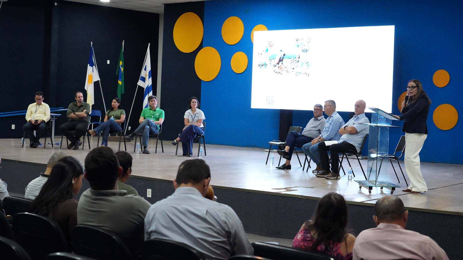 A imagem mostra várias pessoas sentadas em cadeiras no palco, enquanto outras assistem na plateia. A foto foi tirada no auditório da escola estadual Elizangela Glória Cardoso, em Palmas, durante o Seminário de resíduos sólidos. 