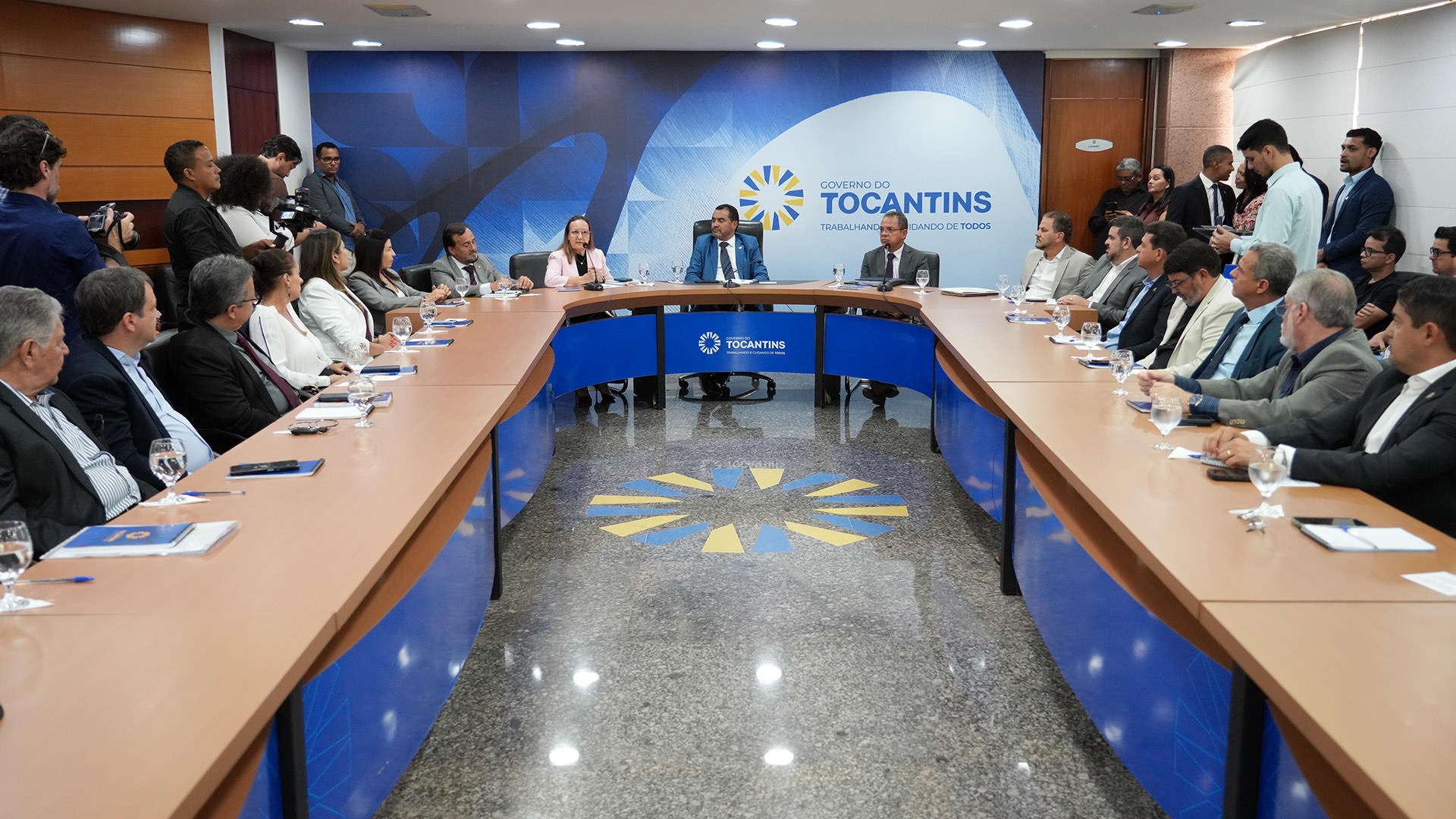 A imagem mostra um grupo de autoridades sentadas ao redor de uma grande mesa, na sala de reuniões do Palácio Araguaia, durante o lançamento do Refis 2025 do Governo do Estado. Os participantes estão vestidos com trajes formais, como ternos e vestidos.