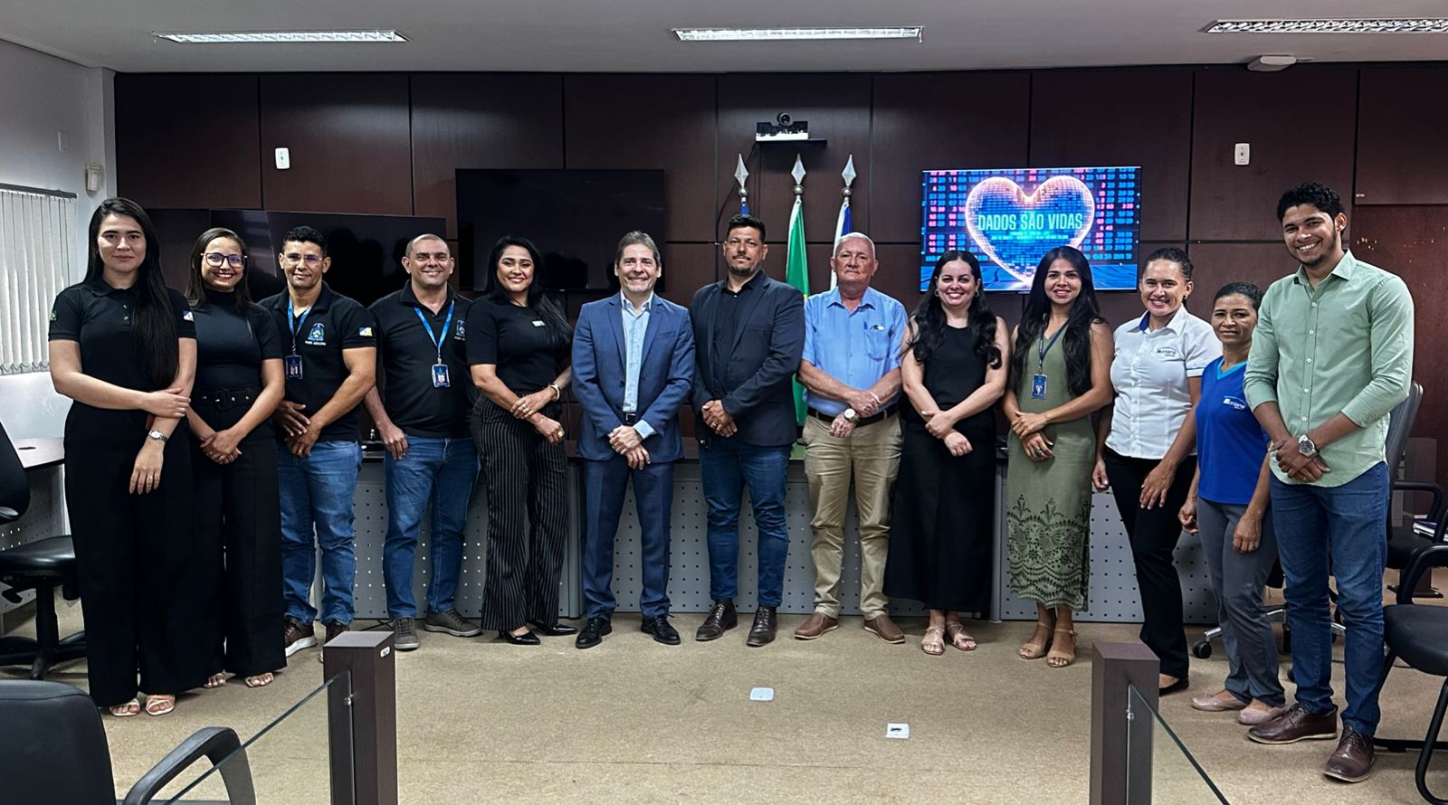 Foto institucional com 15 pessoas em pé, lado a lado, sorrindo para a câmera. Ao fundo, bandeiras do Brasil e Tocantins e uma tela com o logo do projeto “Dados São Vidas”. O grupo está em ambiente formal, com roupas sociais e crachás, em uma sala de reuniões do Judiciário.